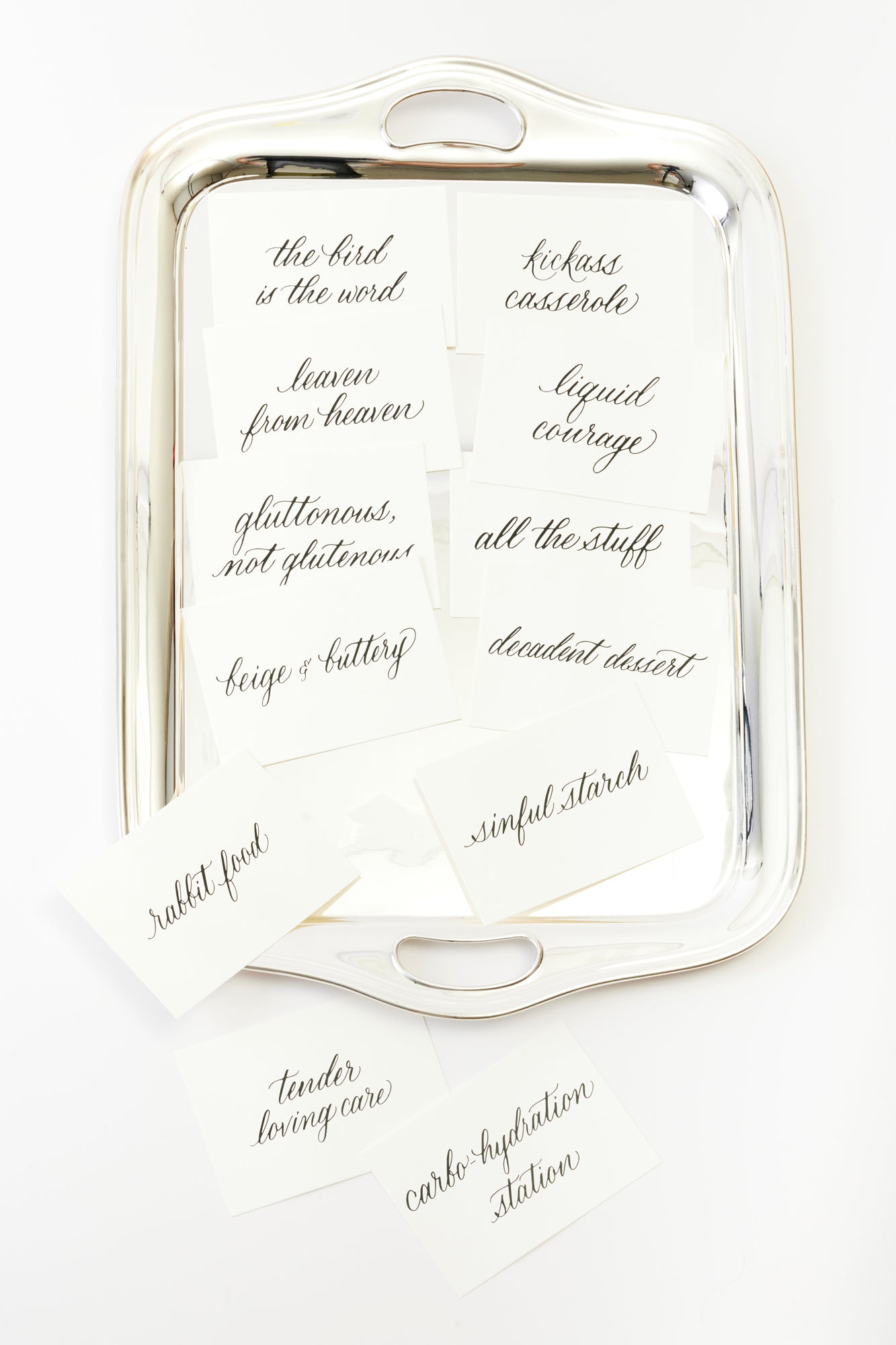 A polished silver tray adorned with elegant buffet label cards featuring whimsical and stylish calligraphy. Phrases like ‘the bird is the word’ and ‘kickass casserole’ bring a playful yet luxurious touch to the dining experience, perfect for a chic, creative event.