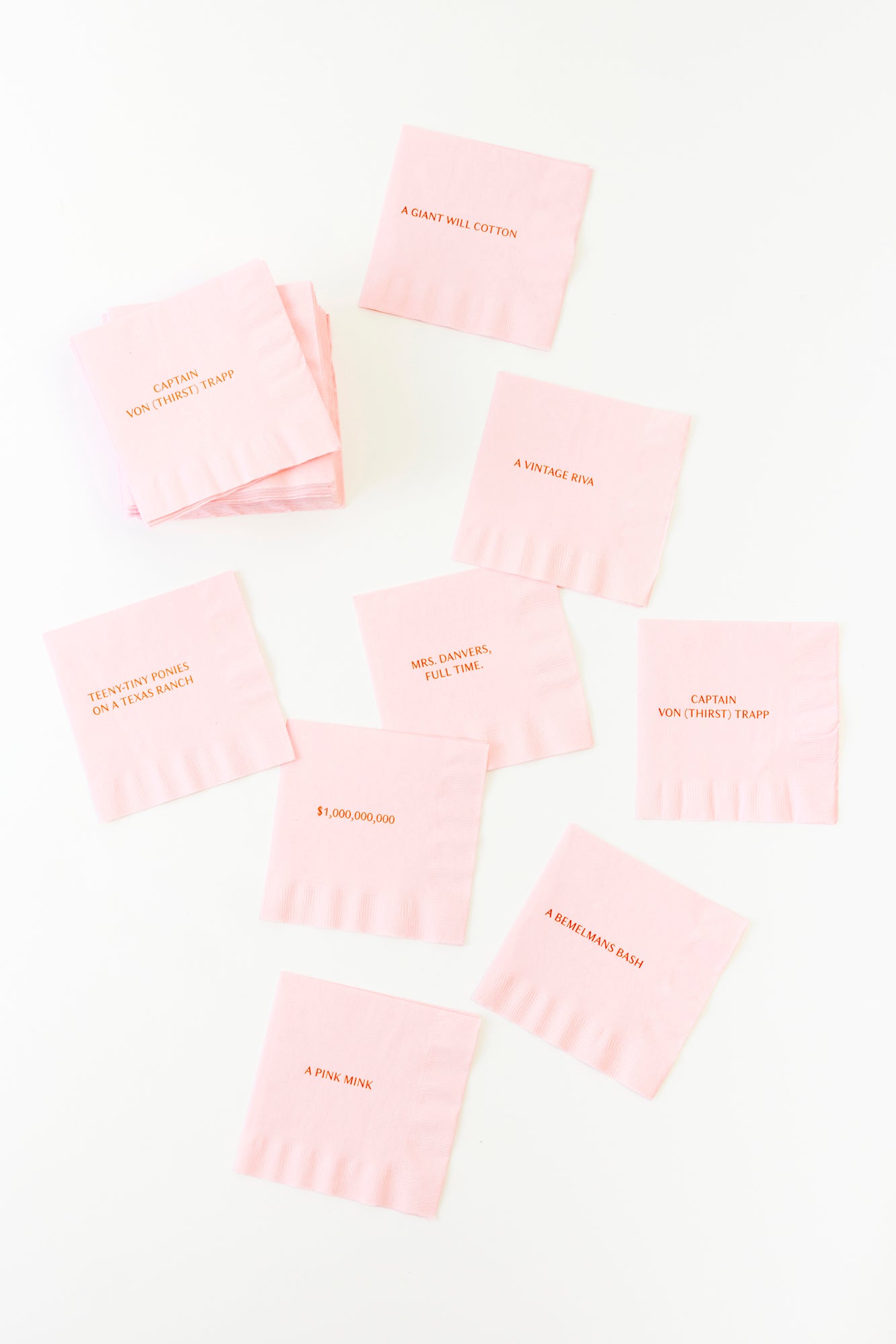Pink paper cocktail napkins scattered across a white background. Each napkin has red text with a variety of Christmas wishlist ideas. Set of 64 includes 8 count of 8 sayings/wishes.
