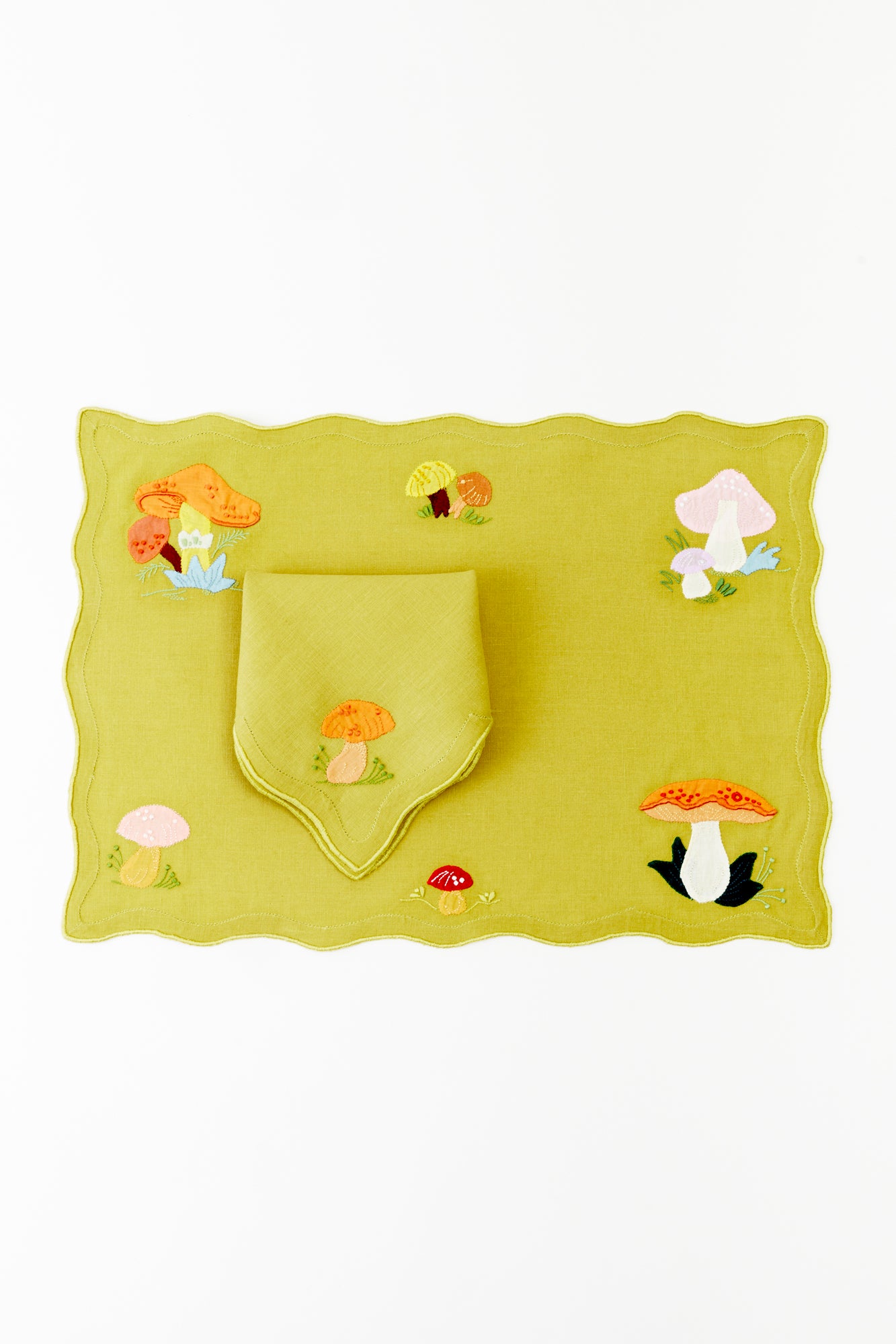 Whimsical lime green linen placemat and matching napkin set, adorned with embroidered mushrooms in shades of orange, pink, and yellow. Delicate wavy edges and playful nature-inspired designs evoke a charming woodland aesthetic, perfect for adding a playful yet chic touch to any table setting.