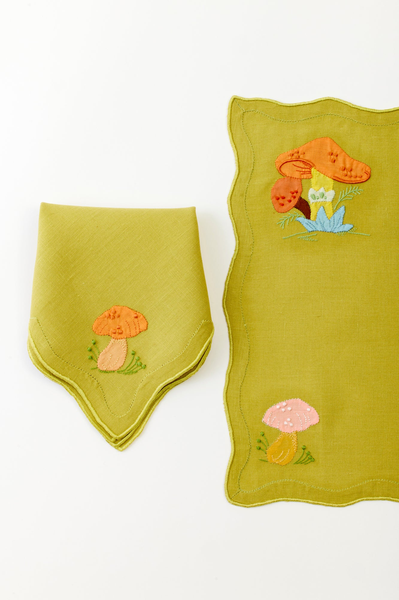 Close-up of an embroidered linen placemat and folded napkin in lime green, showcasing intricately stitched mushrooms in soft pink and orange hues. The playful mushroom embroidery and wavy edge detail enhance the charming and whimsical design, ideal for a trendy, woodland-inspired table setting.