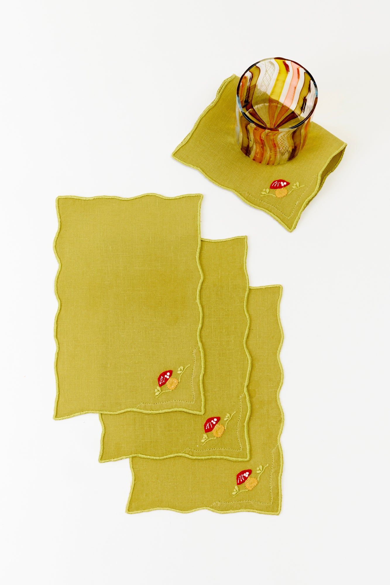 Set of whimsical lime green linen cocktail napkins with embroidered red and white mushrooms in the corner, featuring delicate wavy edges. A stylish amber-toned glass rests atop one napkin, adding a touch of organic elegance to this nature-inspired table accessory, perfect for elevating any forest-themed or rustic gathering.
