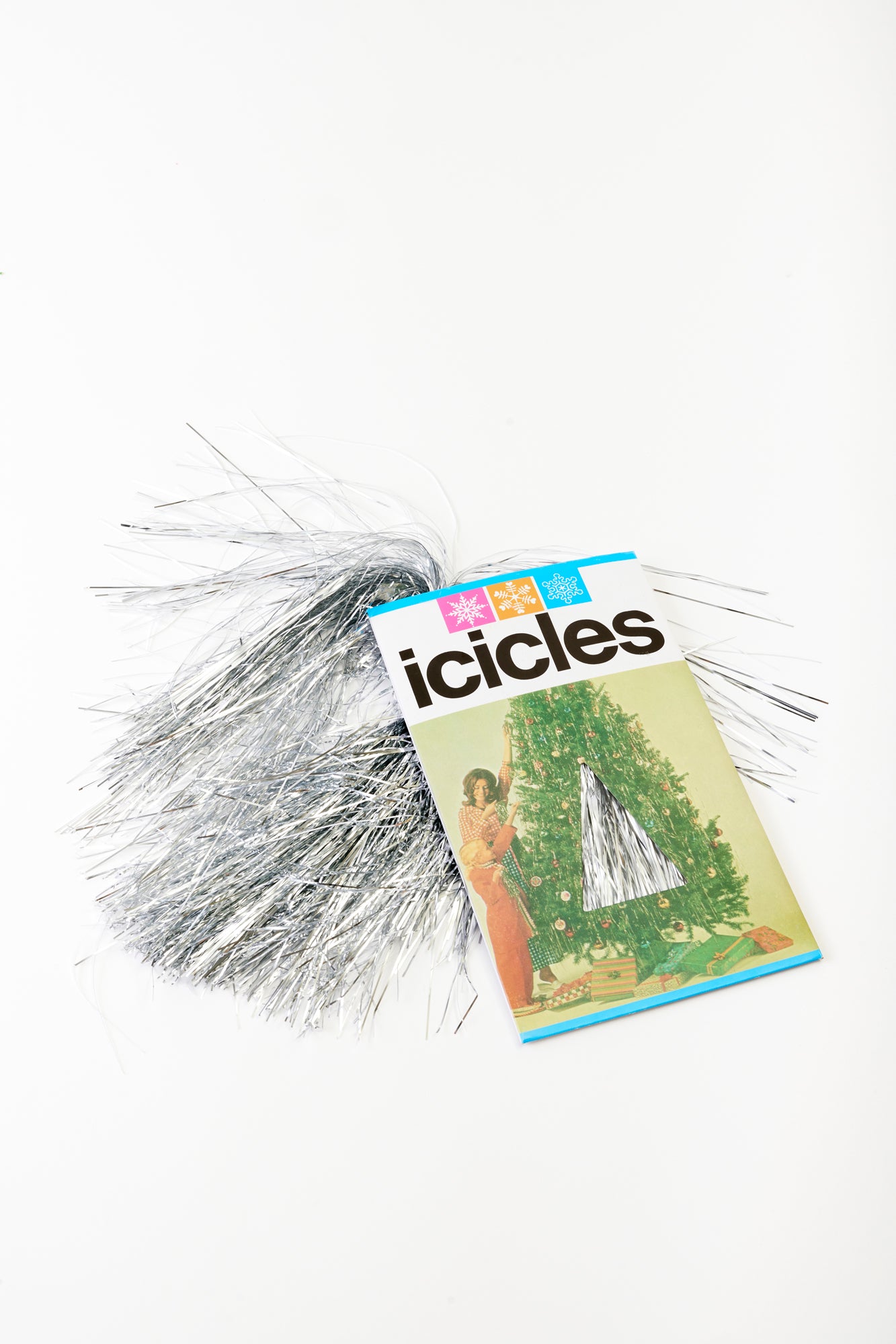 Vintage-inspired holiday icicles tinsel, shimmering in silver, spilling out from a retro-style package featuring a nostalgic Christmas scene. Perfect for adding a touch of classic sparkle and glamour to your festive decor.