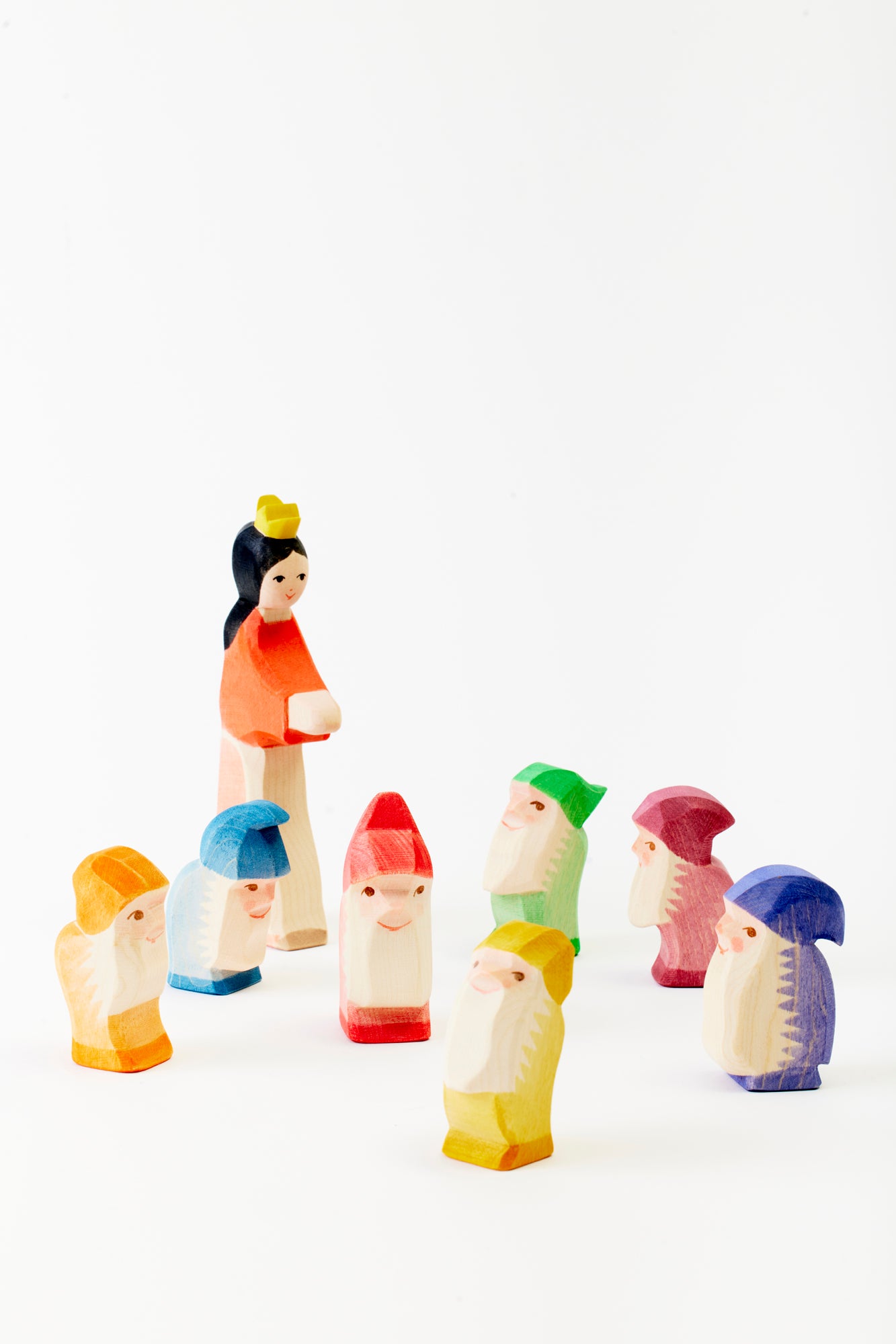 A set of small, colorful wooden figurines arranged in a semicircle. The figurines include a taller Snow White figure wearing an orange top and beige skirt with a yellow crown, standing in the center, and seven shorter dwarves, each wearing different colored hats and tunics in vibrant hues like red, green, blue, yellow, and purple. The figurines are hand-carved with simple facial features, capturing a playful, minimalistic style.