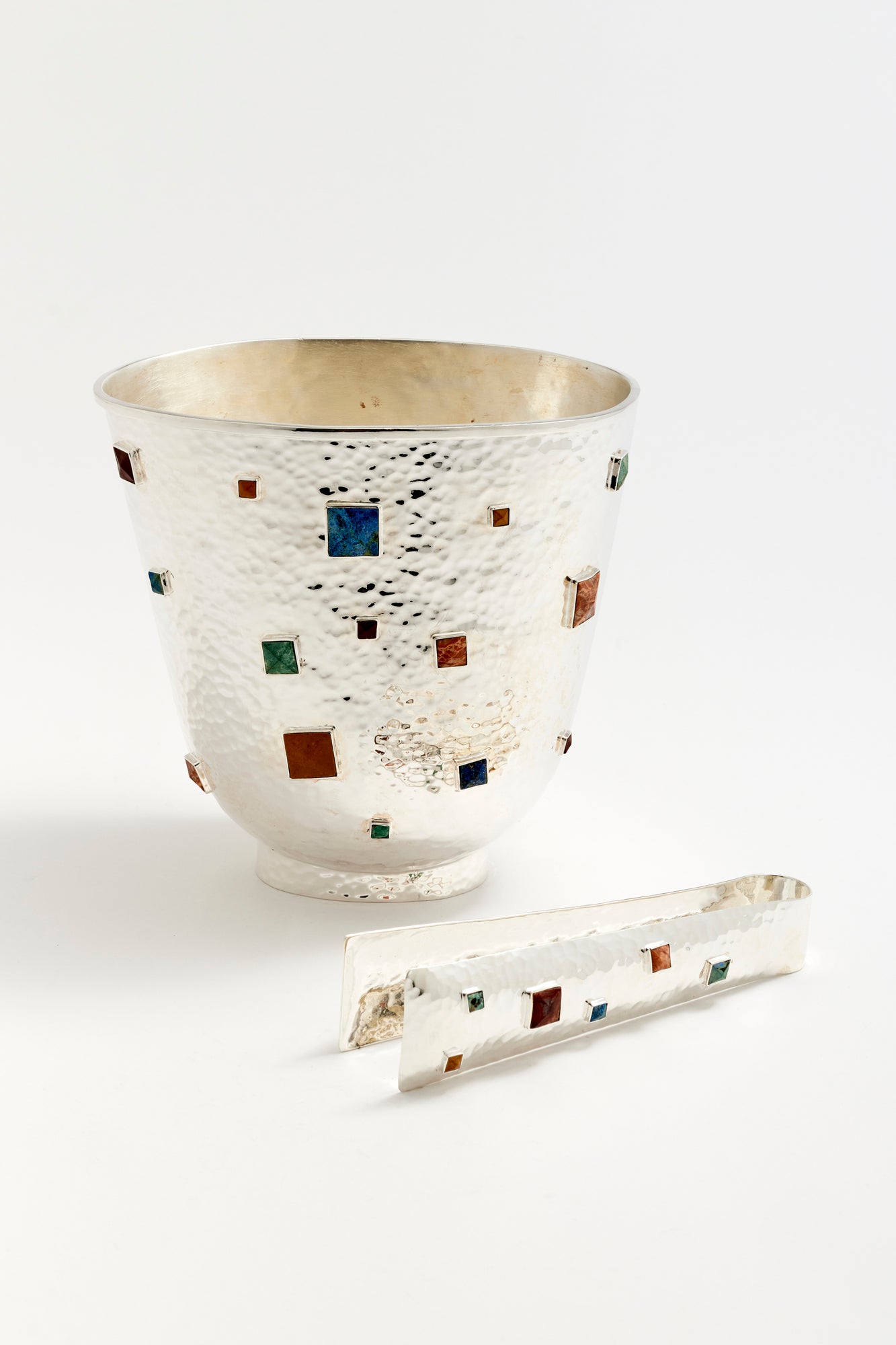 Elegant hammered silver ice bucket with a modern, artisanal design, featuring inlaid semi-precious stones in a variety of rich colors and geometric shapes. Paired with matching silver tongs, this sophisticated piece adds a luxurious touch to any bar or entertaining space, perfect for elevating your cocktail presentation.