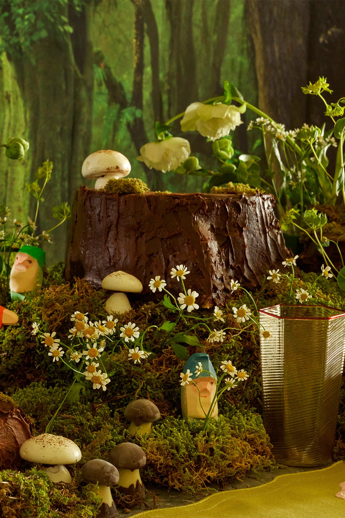 A close-up of a magical woodland tablescape featuring moss-covered surfaces, whimsical wooden figurines, and white and brown mushrooms nestled amongst delicate wildflowers. The scene is anchored by a tree stump centerpiece, creating an enchanting forest-inspired ambiance, while a sleek, modern glass cup adds a subtle contrast to the rustic, nature-themed décor.