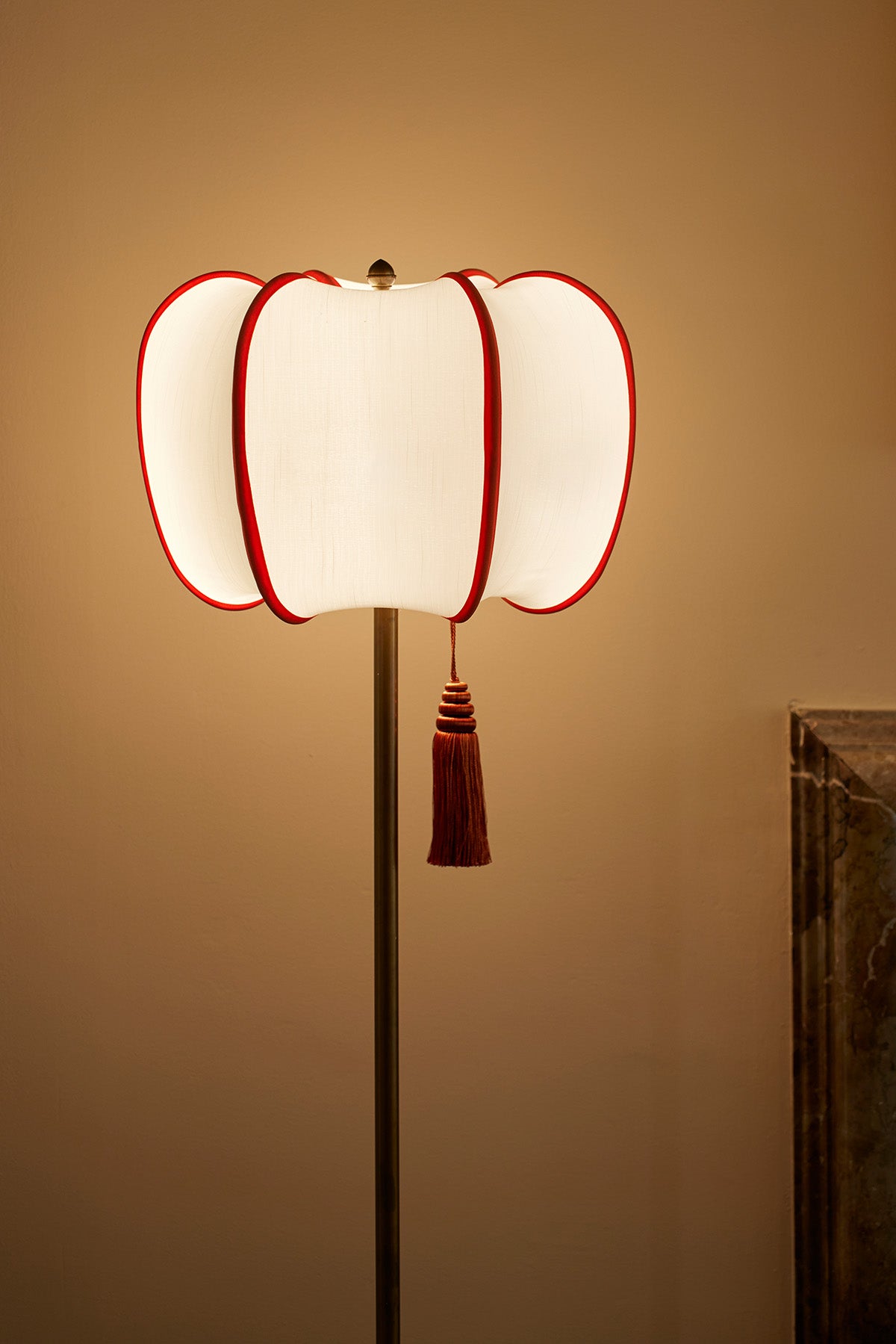 An elegant, vintage-inspired, pumpkin shaped floor lamp with a cream-colored fabric shade outlined in bold red trim. The subtle tassel detail adds a touch of classic luxury, casting a warm, sophisticated glow perfect for high-style interiors.