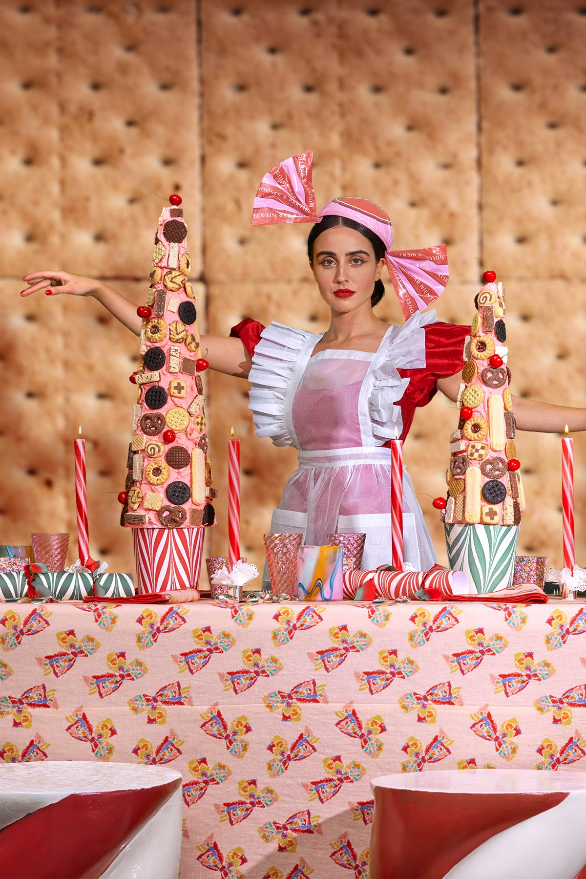 A woman dressed as a whimsical pastry chef stands behind a festive dessert table, wearing a red and white ruffled apron with a matching candy headpiece. The table is decorated with candy cane-striped candles, colorful cups, and towering dessert trees adorned with cookies and sweets. The background features a giant graham cracker wall, and the tablecloth is patterned with vintage holiday imagery, creating a vibrant and playful holiday scene reminiscent of a candy wonderland.
