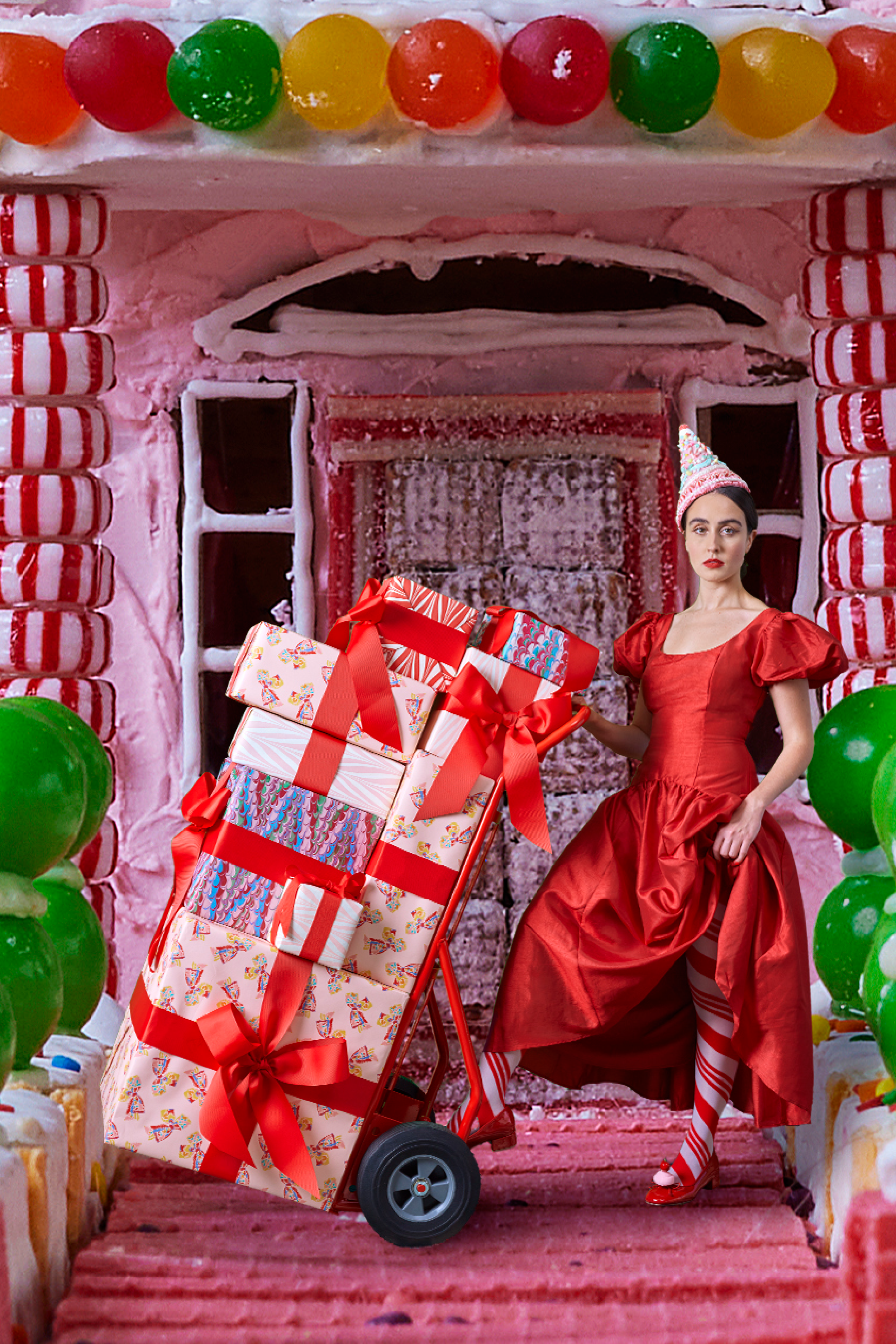 Model in a red dress and candy cane striped stockings pushing a cart filled with wrapped presents, featuring red ribbons and colorful wrapping paper, in front of a whimsical gingerbread-style house.
