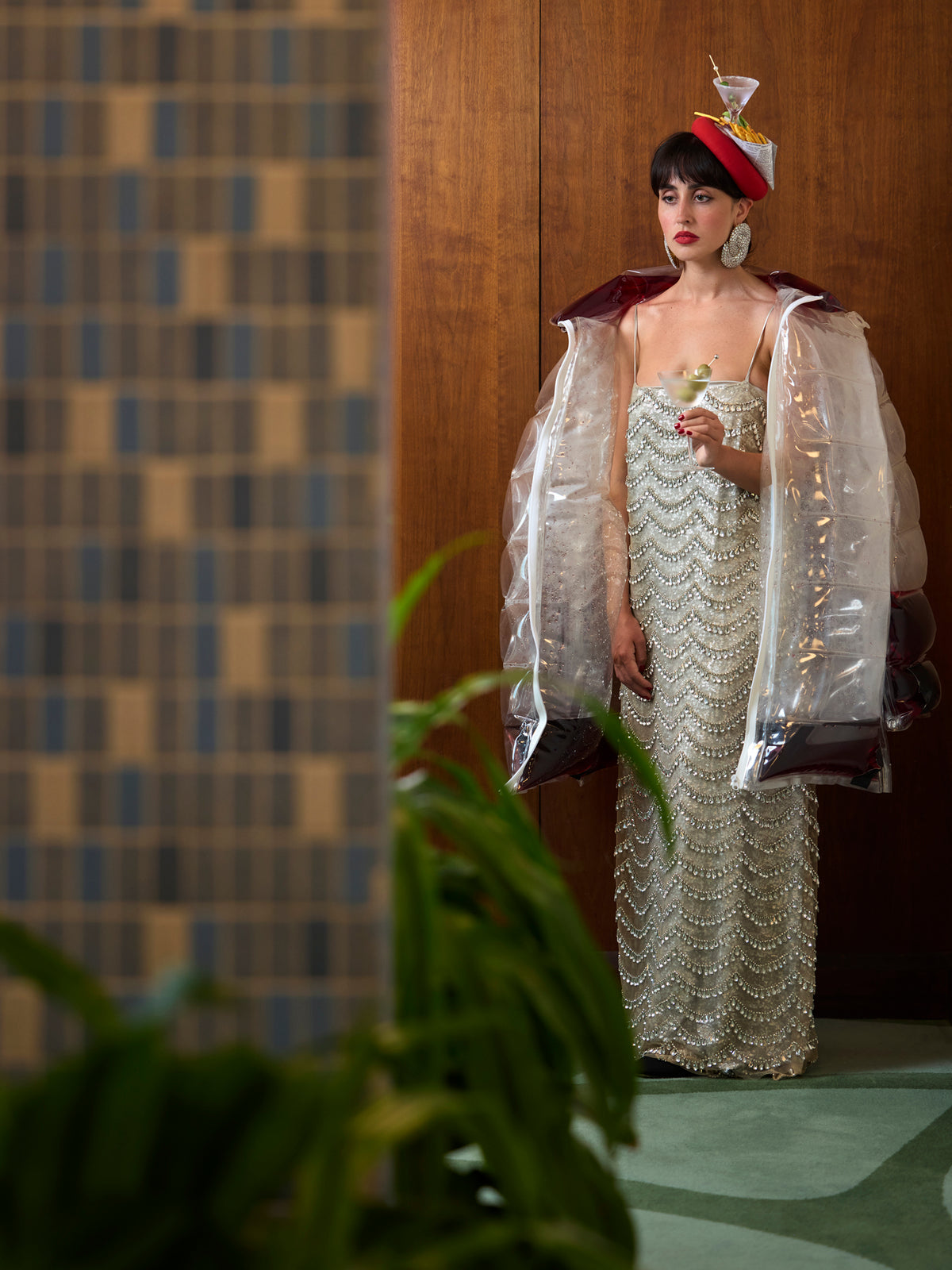 A model stands indoors wearing a long, embellished silver gown with a translucent inflatable puffer coat filled with red wine, wearing a he'd hat with a Martini, Caesar salad, and French fries on top. The is holding a martini with olives in a room with warm red paneling.