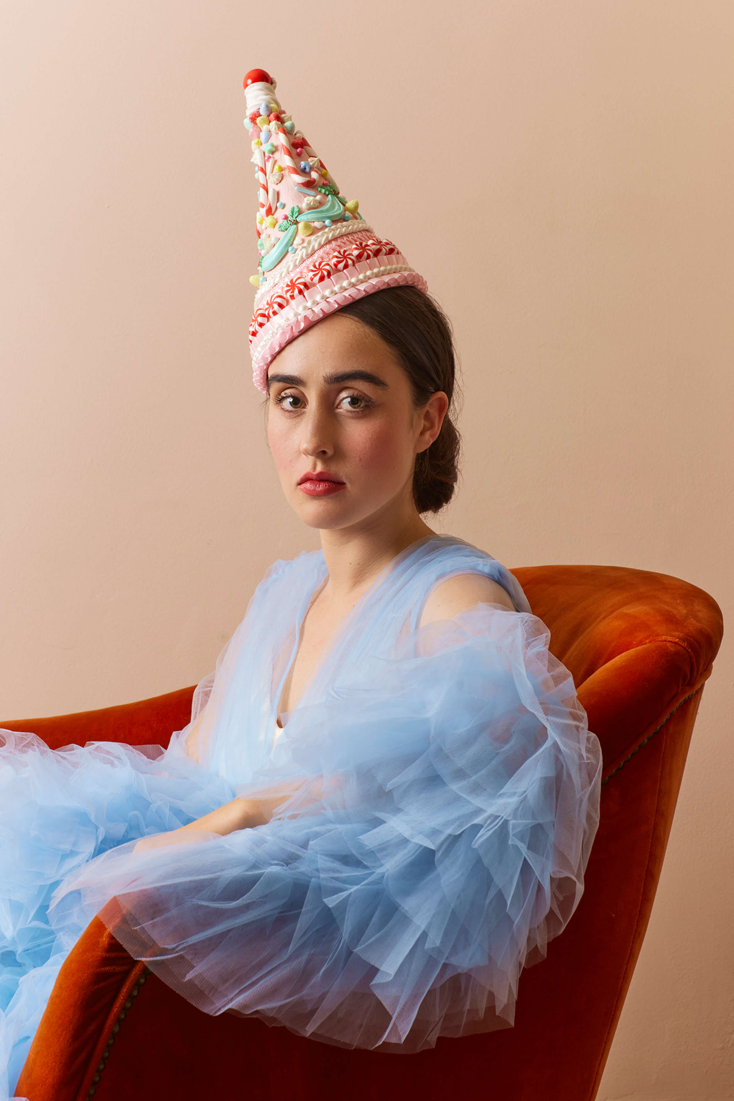 Woman wearing a colorful party hat and light blue dress sitting on an orange chair against a beige background