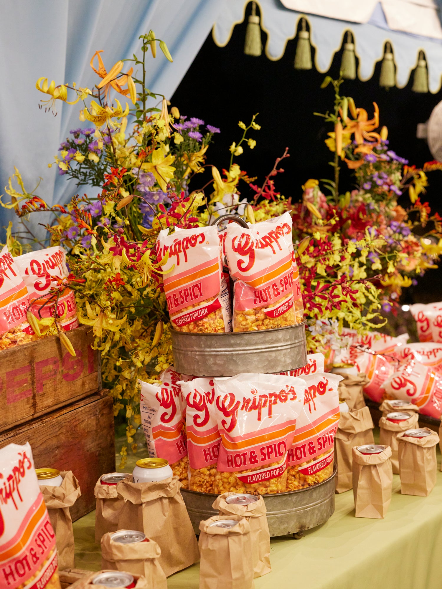 Artfully styled refreshment station featuring vintage-inspired hot and spicy popcorn bags displayed in galvanized metal buckets, surrounded by vibrant wildflower arrangements in wooden crates and craft paper-wrapped beverages – an elevated, nostalgic concession experience for a sophisticated event.