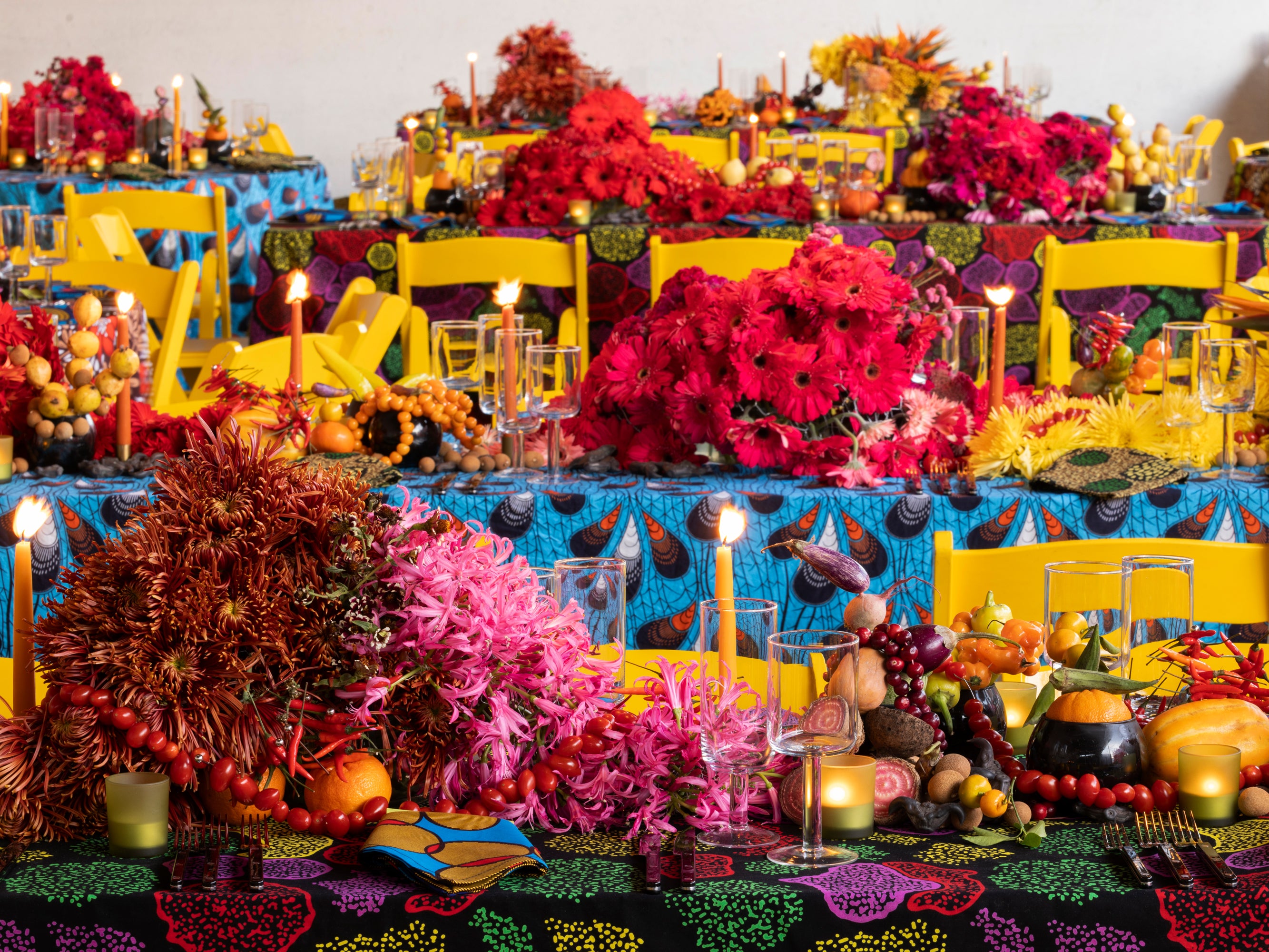 A series of tables set with bright ankara tablecloths, yellow chairs, candelight, and fresh produce tumbling down the centers of each.