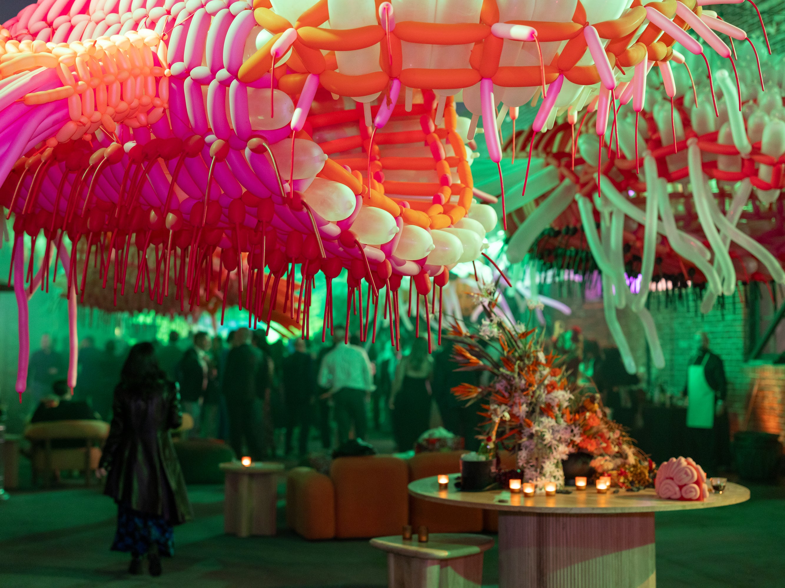 An abstract balloon sculpture hangs over a party filled with people and green mood lighting.