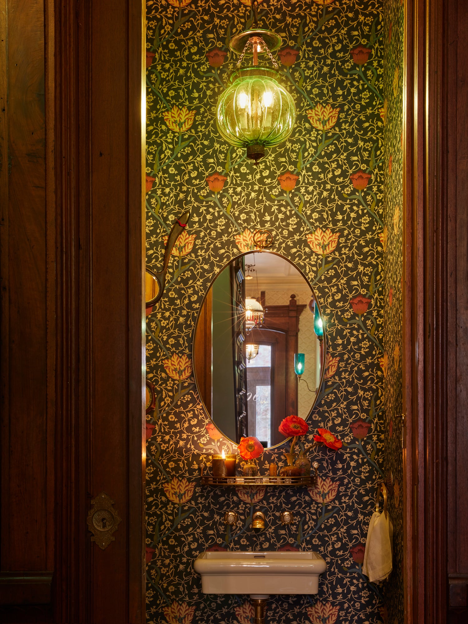 Houses & Parties powder room showcasing rich William Morris-inspired wallpaper in dark green with coral and gold floral motifs. A simple white porcelain sink is mounted beneath an oval brass mirror, with vibrant red poppies and a lit candle creating a dramatic focal point. A vintage-inspired glass pendant light with exposed bulbs illuminates the intimate space, framed by dark wood doorway. This moody jewel-box bathroom exemplifies the brand's luxurious yet irreverent approach to small spaces.