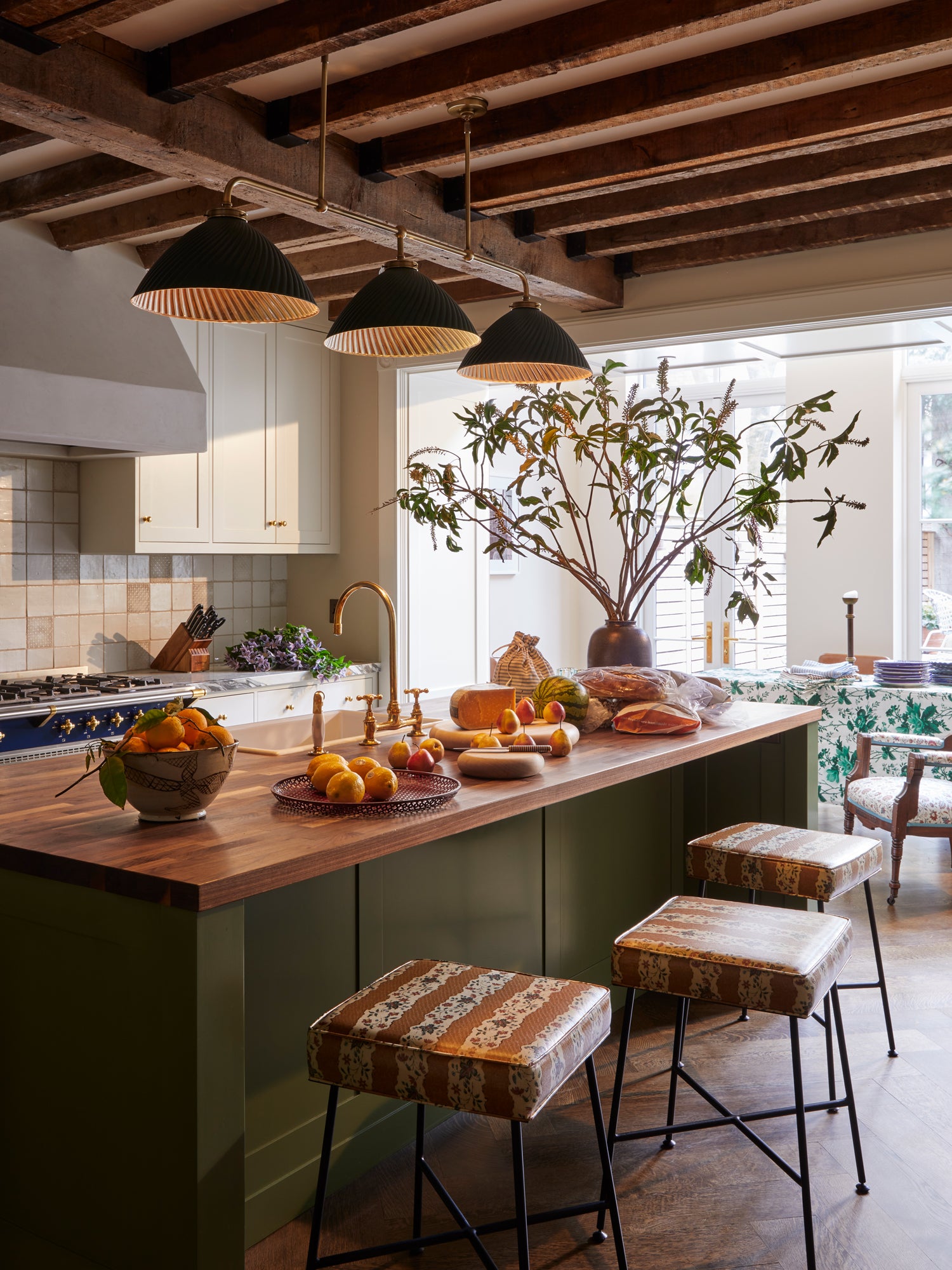 Houses & Parties rustic-modern kitchen featuring exposed wooden ceiling beams above an olive-green island with butcher block countertop abundant with fresh produce. Three black pendants with gold interiors illuminate the space while vintage-patterned bar stools add character. White cabinetry, handmade tile backsplash, and brass fixtures create warmth, with a statement botanical arrangement framed by a bright window.