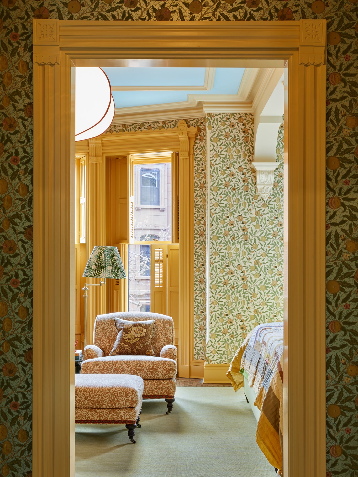 Bedroom viewed through a doorway framed by rich fruit-patterned William Morris wallpaper. Inside, a harmonious layering of patterns continues with leafy green wallpaper against warm wooden moldings and a pale blue skylight. A cozy reading nook features a coral-patterned armchair and matching ottoman beside a floor lamp with botanical shade. The corner of a bed with patterned throw is visible, exemplifying the brand's sophisticated approach to mixing heritage prints with classical architectural elements.