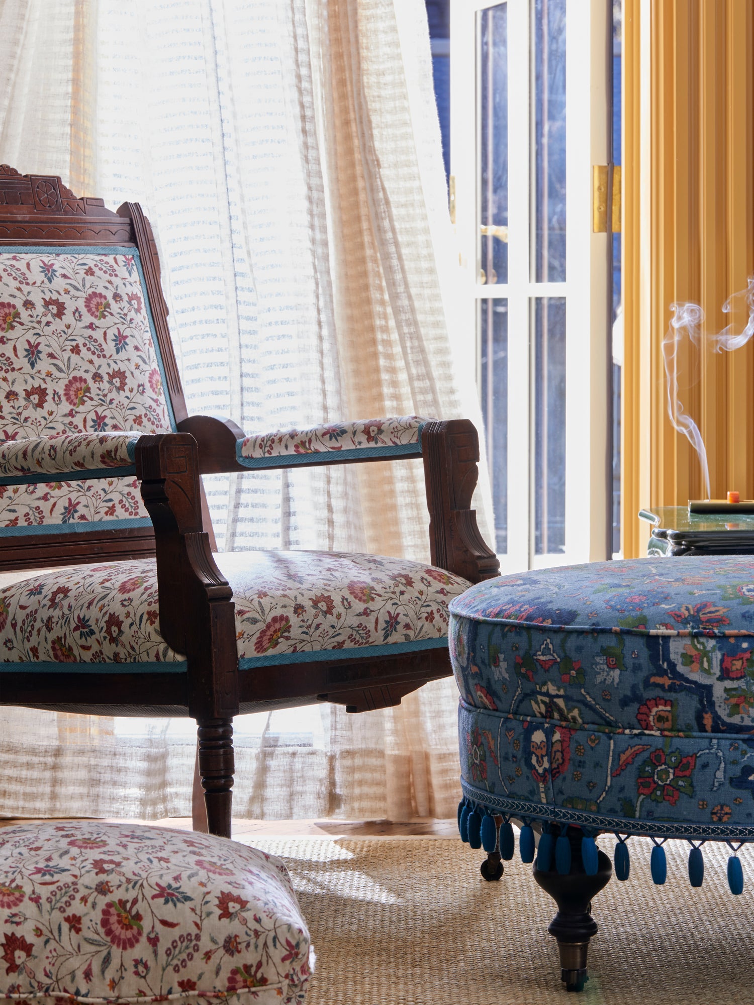 Houses & Parties sitting area featuring an antique wooden chair upholstered in delicate floral fabric with blue piping. Beside it stands a distinctive blue ottoman with vibrant folkloric textile covering and playful tasseled trim. Gauzy linen curtains filter sunlight through French doors, while wisps of incense smoke curl upward from the side table. The contrasting patterns and unexpected textural elements exemplify the brand's signature blend of traditional forms with irreverent, eclectic styling.