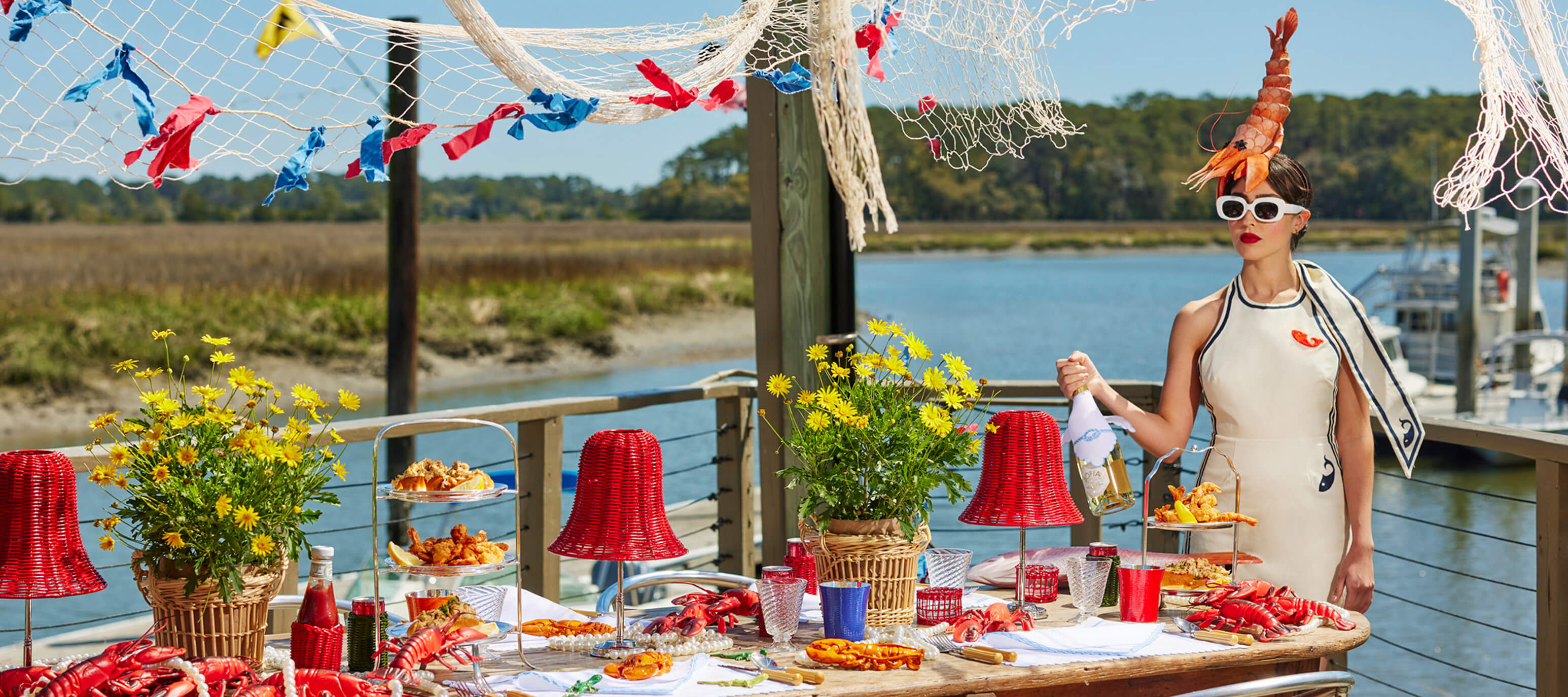 A woman in a shrimp headpiece stands by a seaside table set with lobster, red lamps, and nautical decor, holding a bottle of champagne.
