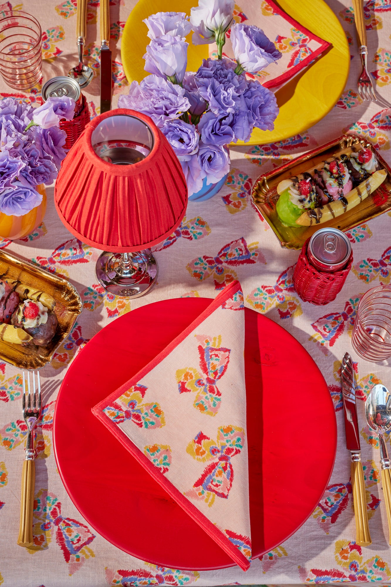 Table setting featuring a pink tablecloth with a printed pattern of a vintage-inspired rainbow bow. Atop the table are colorful place settings, red shaded table lamps, bamboo flatware, red wicker can koozies, and fresh purple flowers in blown glass vases.