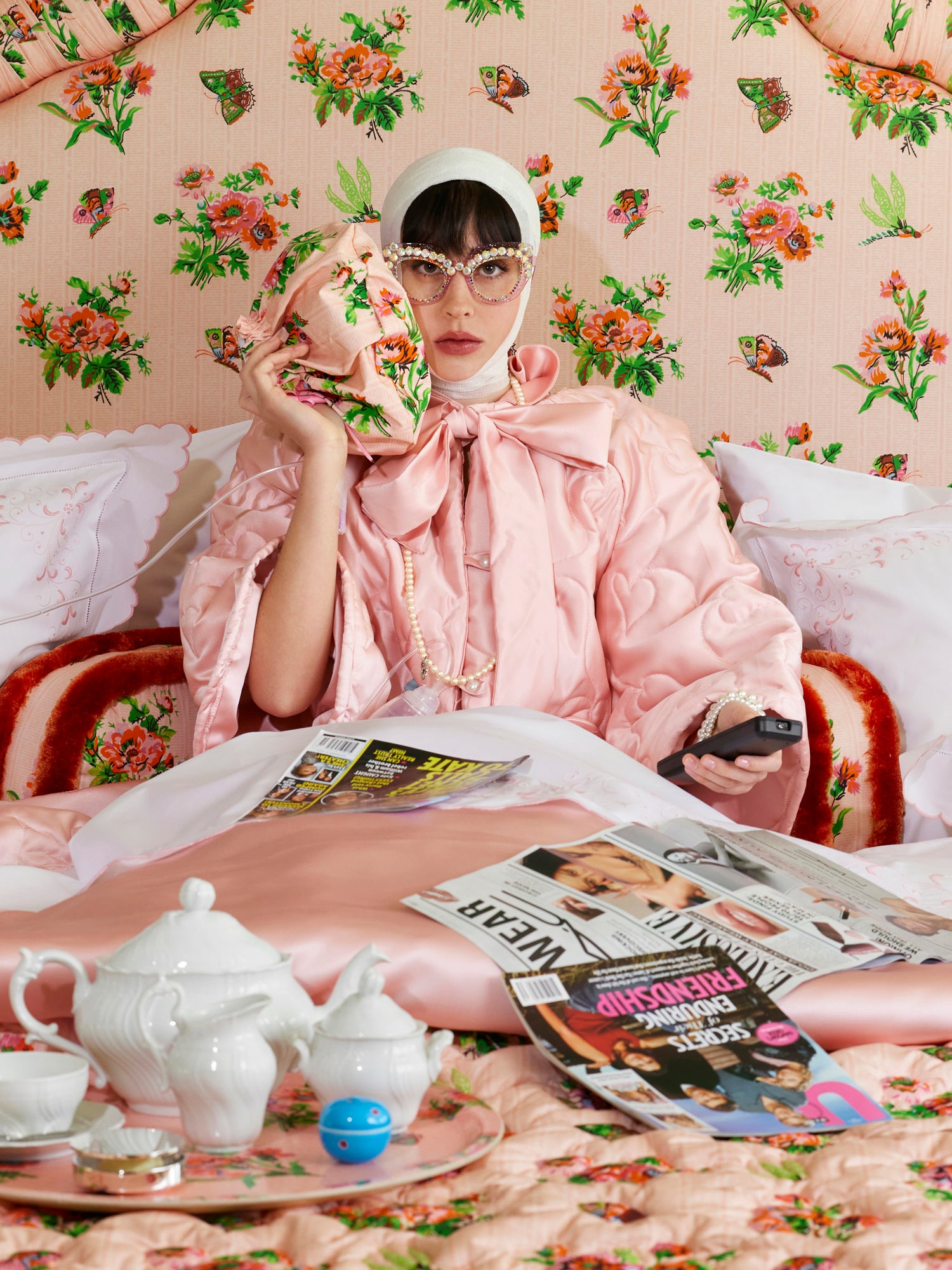 Woman in a pink quilted bed jacket, sitting against a floral chintz headboard and holding a pink ice pack to her face.