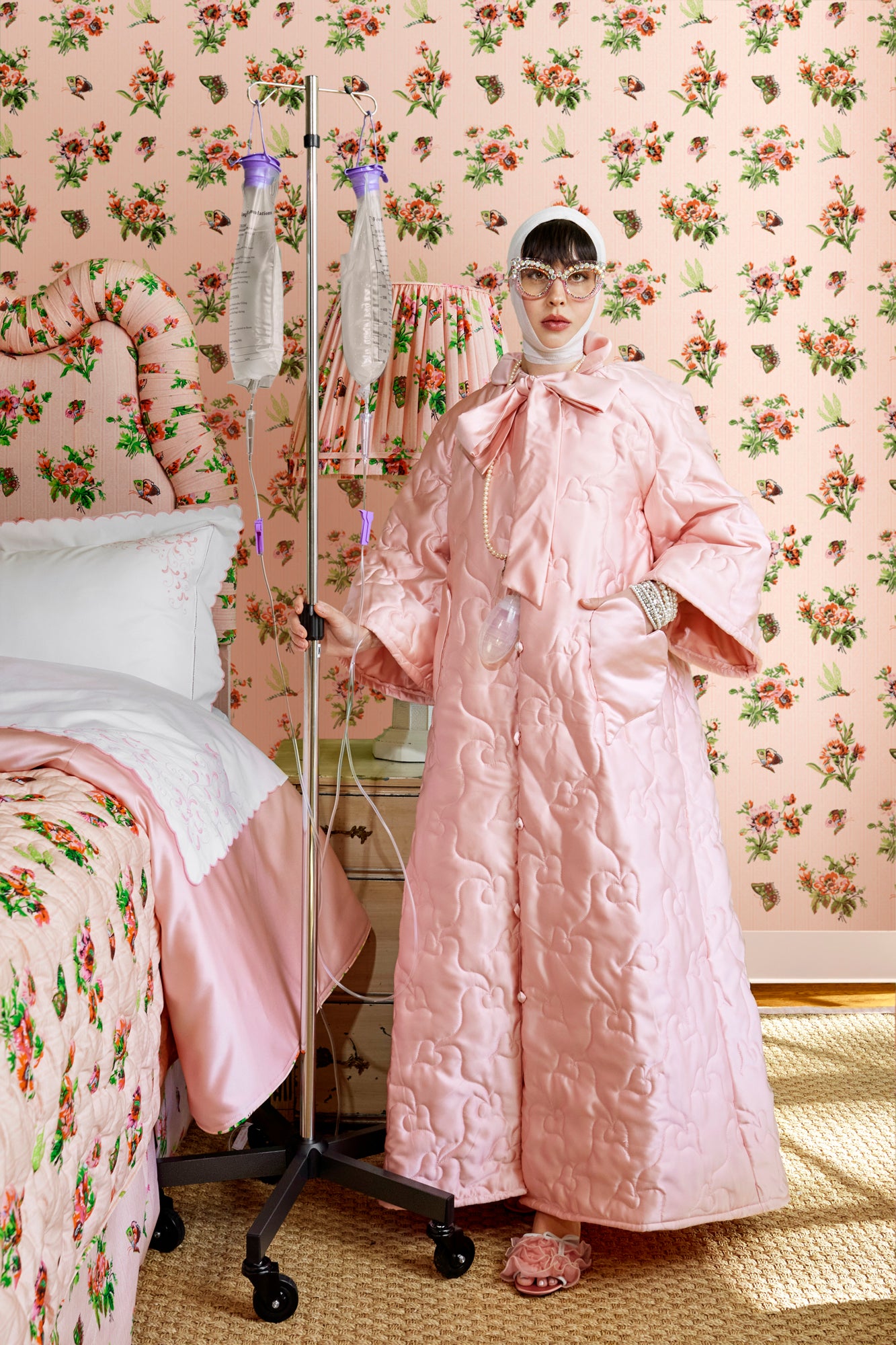 Person wearing a pink quilted house coat in a room with pink chintz wallpaper, quilt, and headboard.