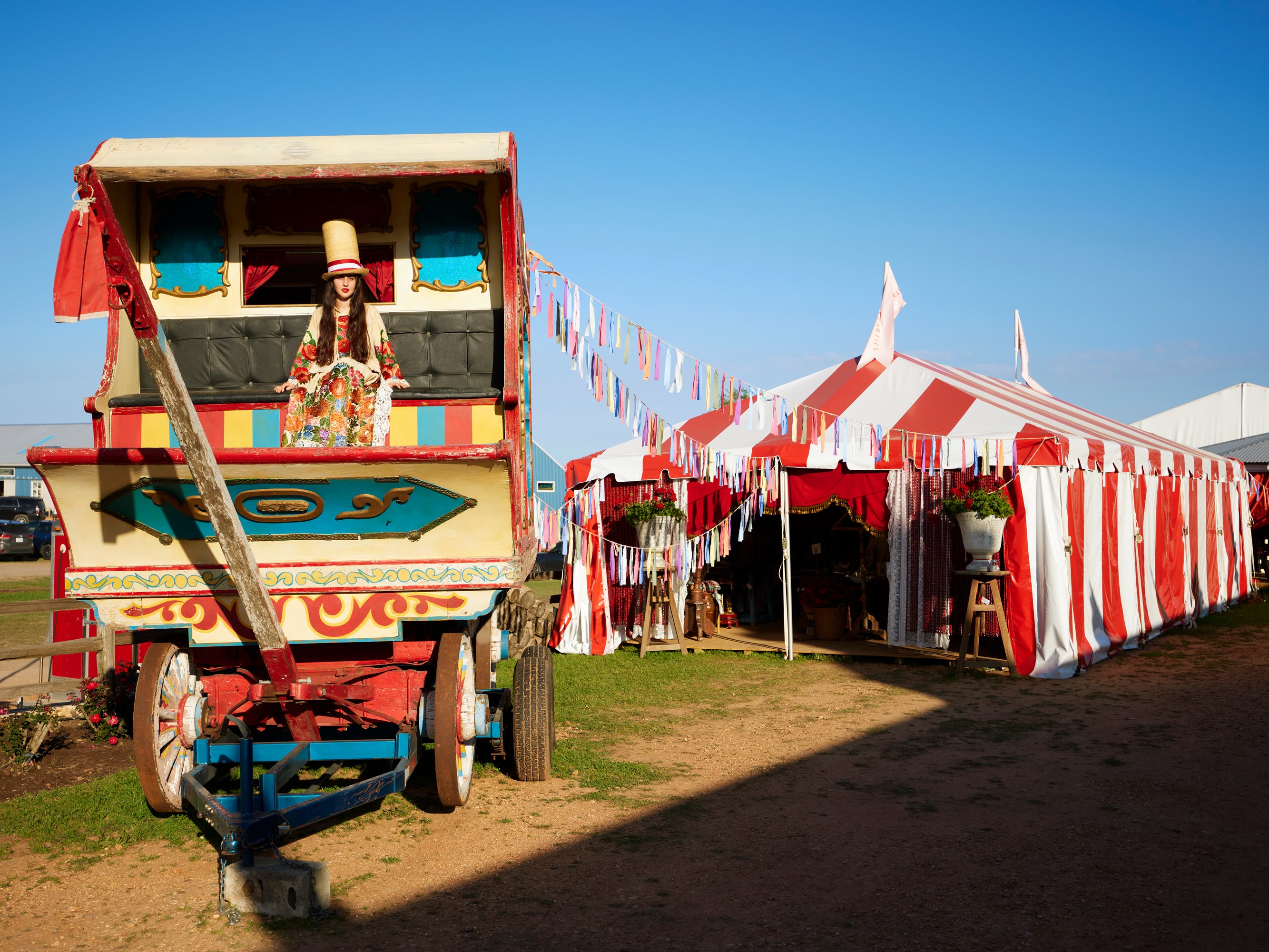 Theatrical event installation featuring a colorful vintage gypsy caravan with a costumed woman in a top hat, connected by festive bunting to a red-and-white striped tent pavilion – a whimsical carnival-inspired setting for an extraordinary luxury celebration.