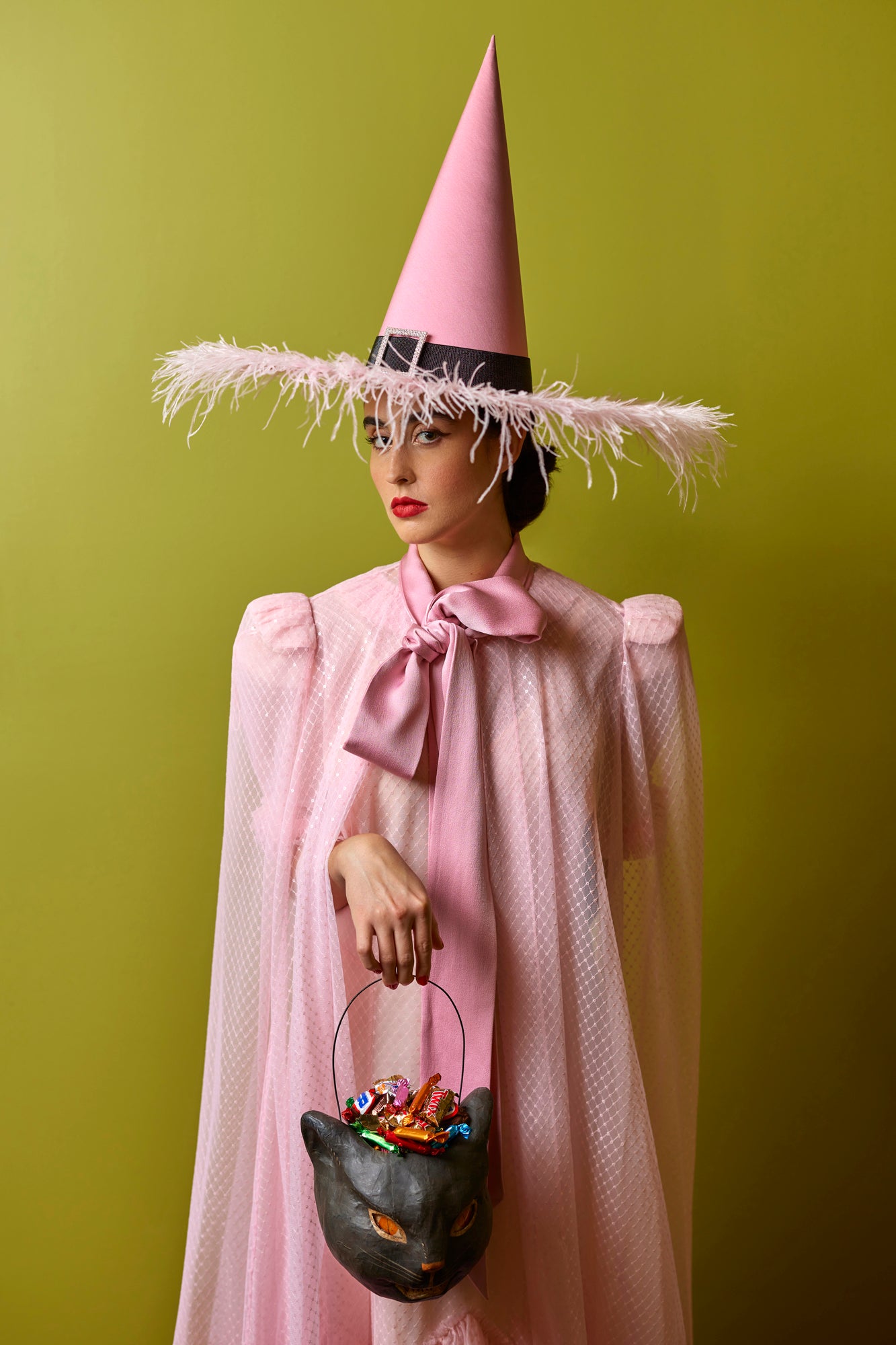 A woman dressed as a stylish pink witch, wearing a tall, pointed pink hat with a wide brim adorned with feather trim. She is also holding a black cat-shaped bucket.