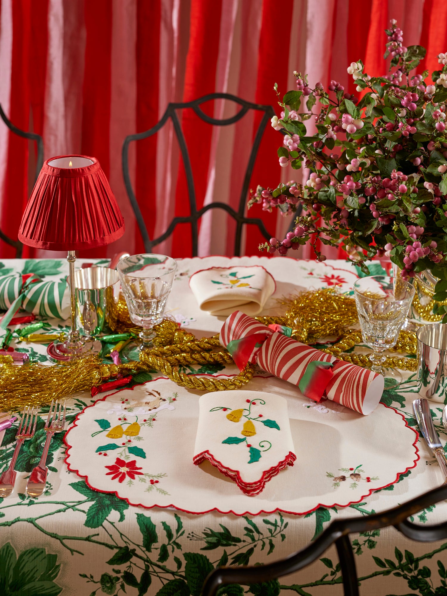 Christmas themed table setting featuring silver tumbler, crystal goblet, silver table lamp with red shade, linen placemat and napkin with French embroidered appliqué, fresh florals, and marbled paper party crackers.
