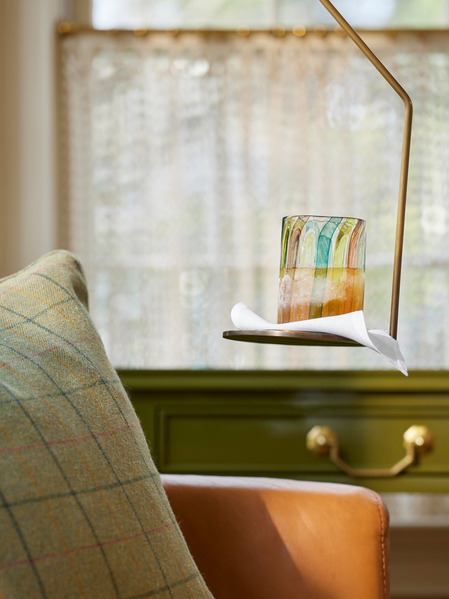 Houses & Parties intimate vignette featuring a brass reading lamp extending over a drink on a small side table. The colorful tumbler with watercolor-like stripes in greens, yellows, and oranges sits atop a white napkin. A leather armchair with sage green windowpane plaid cushion partially frames the scene, while an olive green cabinet with brass hardware is softly blurred in the background. Diffused natural light filters through sheer curtains, creating a cozy, sophisticated moment of repose.