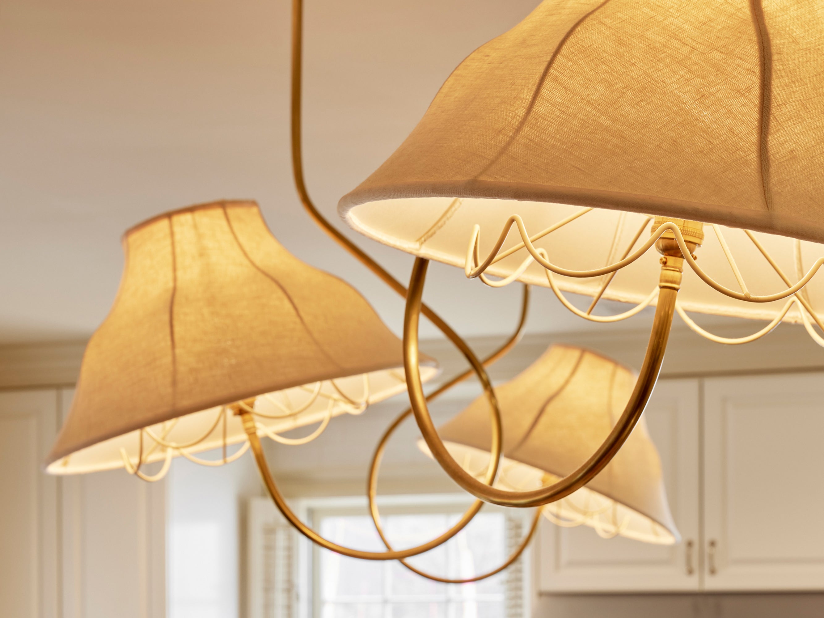 Elegant lighting fixture featuring scalloped linen shades suspended from an interconnected brass frame, casting a warm, inviting glow – a signature Houses & Parties design choice that introduces sculptural interest and softened illumination.