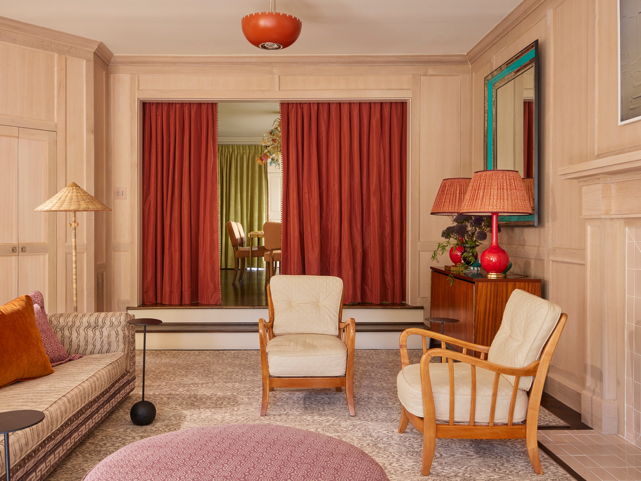 Refined living room featuring cream paneled walls, persimmon silk draperies framing a glimpsed dining space, and mid-century blonde wood armchairs paired with a coral statement lamp – demonstrating Houses & Parties' masterful use of rich color against neutral backgrounds and sophisticated mix of vintage and contemporary furniture.