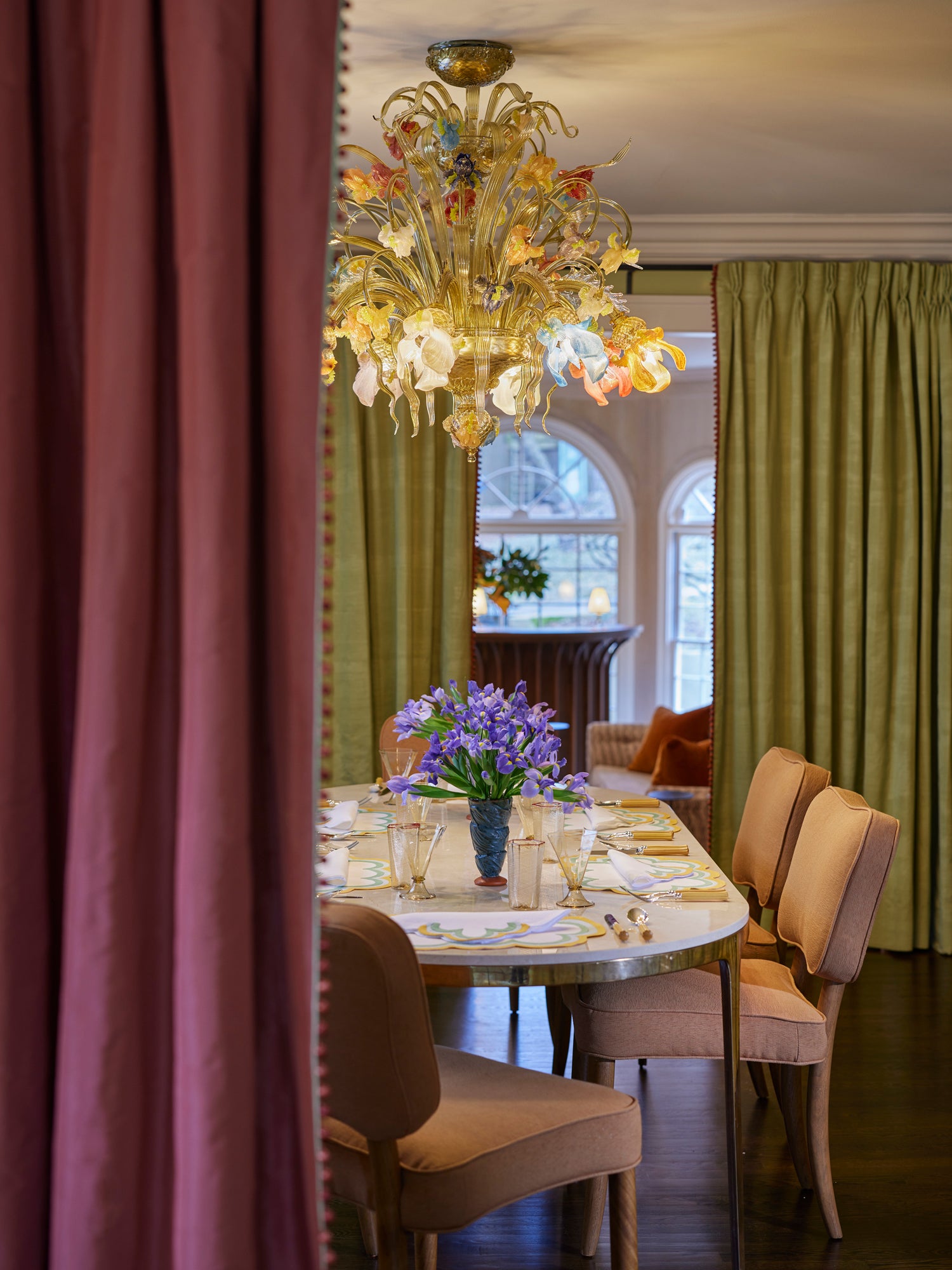 Exquisite dining room featuring a spectacular Venetian glass chandelier above a marble-topped table set with elegant placemats, framed by plum and olive draperies with decorative trim – showcasing Houses & Parties' talent for creating richly layered, classically-inspired spaces that combine antique lighting, fresh florals, and complementary textiles for sophisticated entertaining areas.