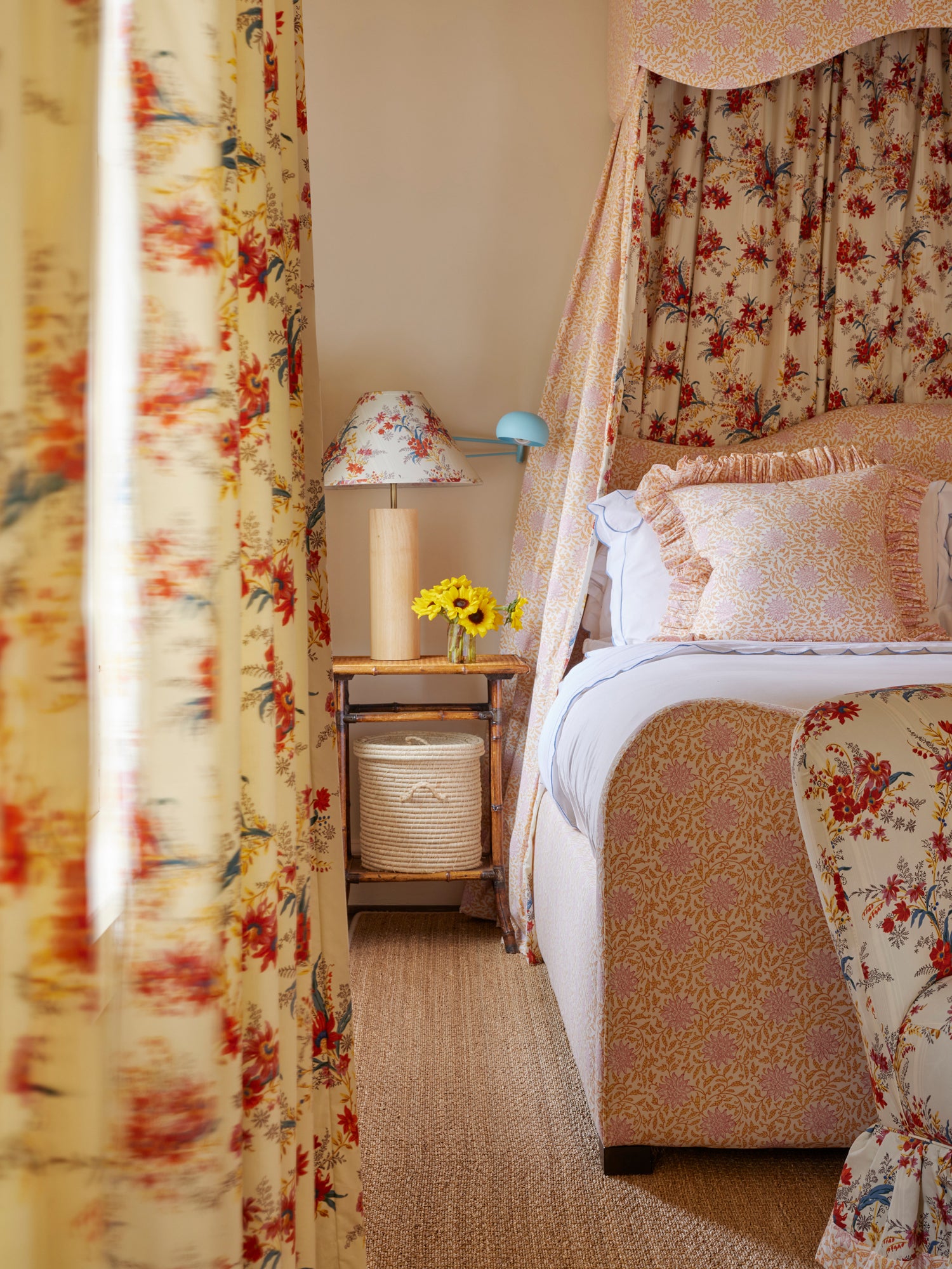 Countryside bedroom featuring a canopy bed draped in complementary floral and leopard-print fabrics in warm cream and red tones. A rustic wooden bedside table holds a minimalist lamp with matching floral shade and cheerful yellow flowers. White linens provide crisp contrast against the patterned textiles, while a woven basket adds natural texture to this luxuriously layered retreat.