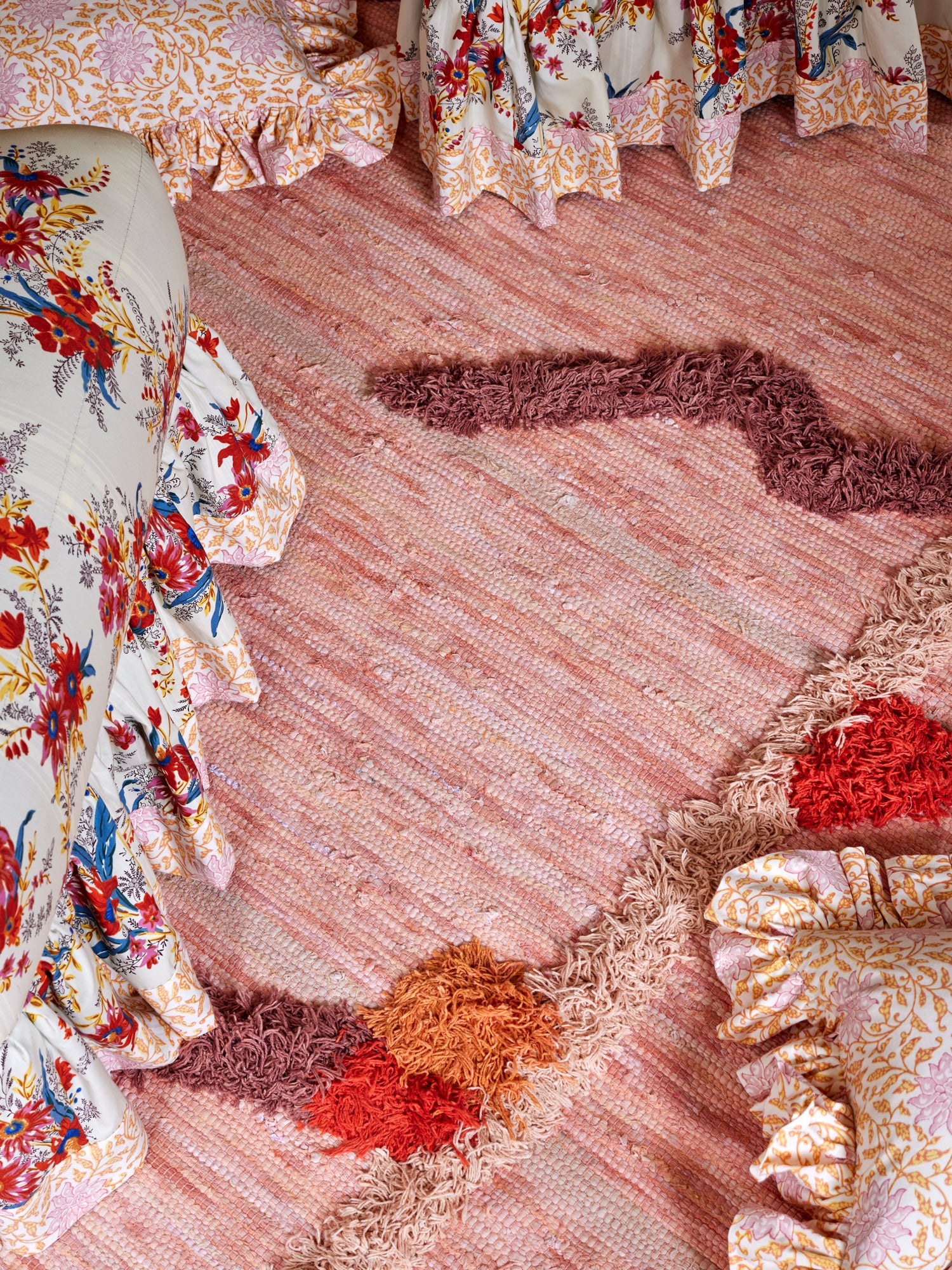 Artfully layered textiles featuring a handcrafted blush area rug with decorative tufted embellishments complemented by vibrant floral fabric with coordinating ruffled trim – showcasing the Houses & Parties commitment to tactile luxury and detailed craftsmanship in their residential and event designs.