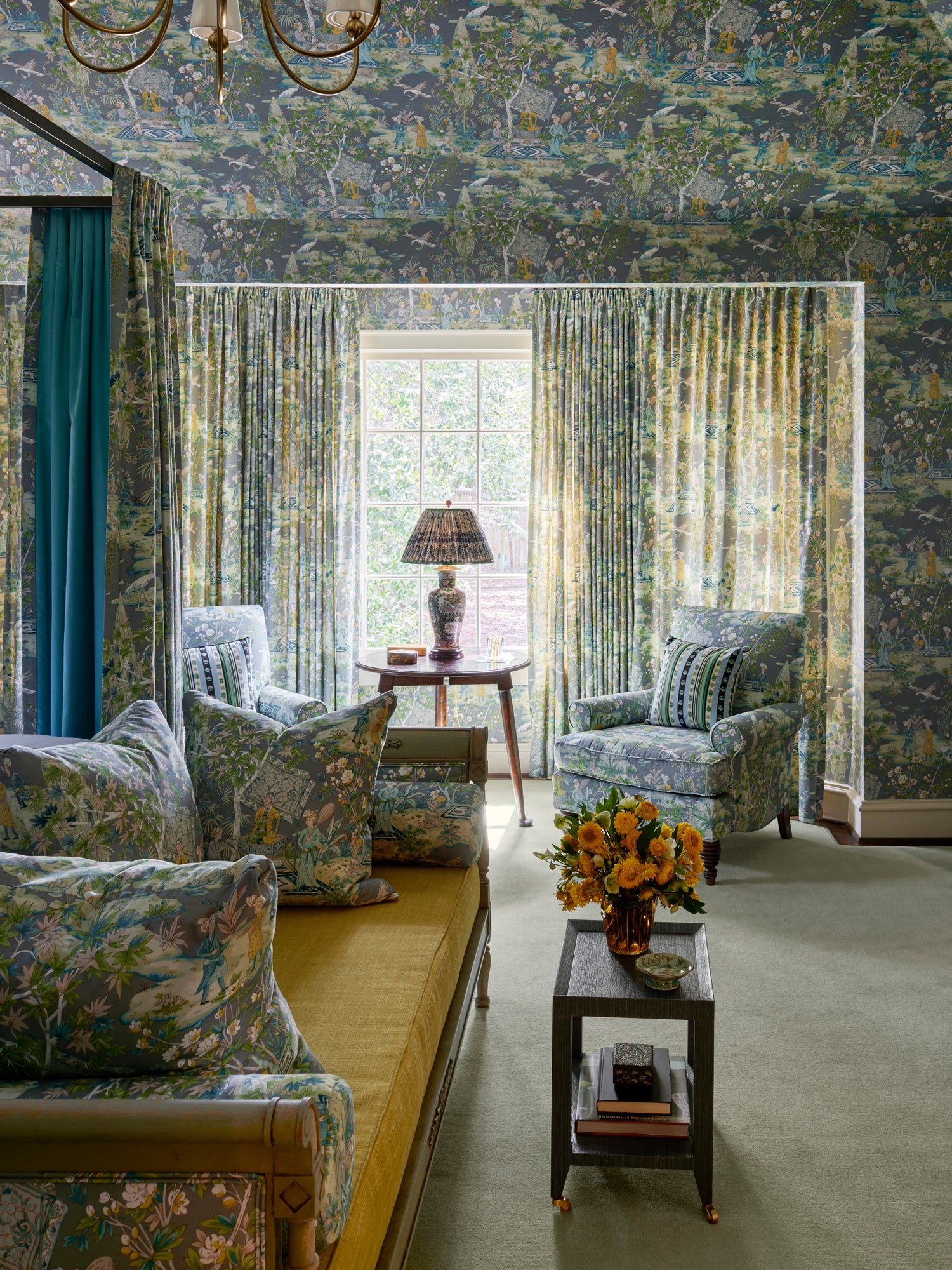 Maximalist living room by Houses & Parties with matching chinoiserie wallpaper, curtains and upholstery in blue-green tones. A mustard daybed contrasts with the botanical pattern, while peach flowers add warmth to this boldly cohesive luxury interior where pattern reigns supreme.