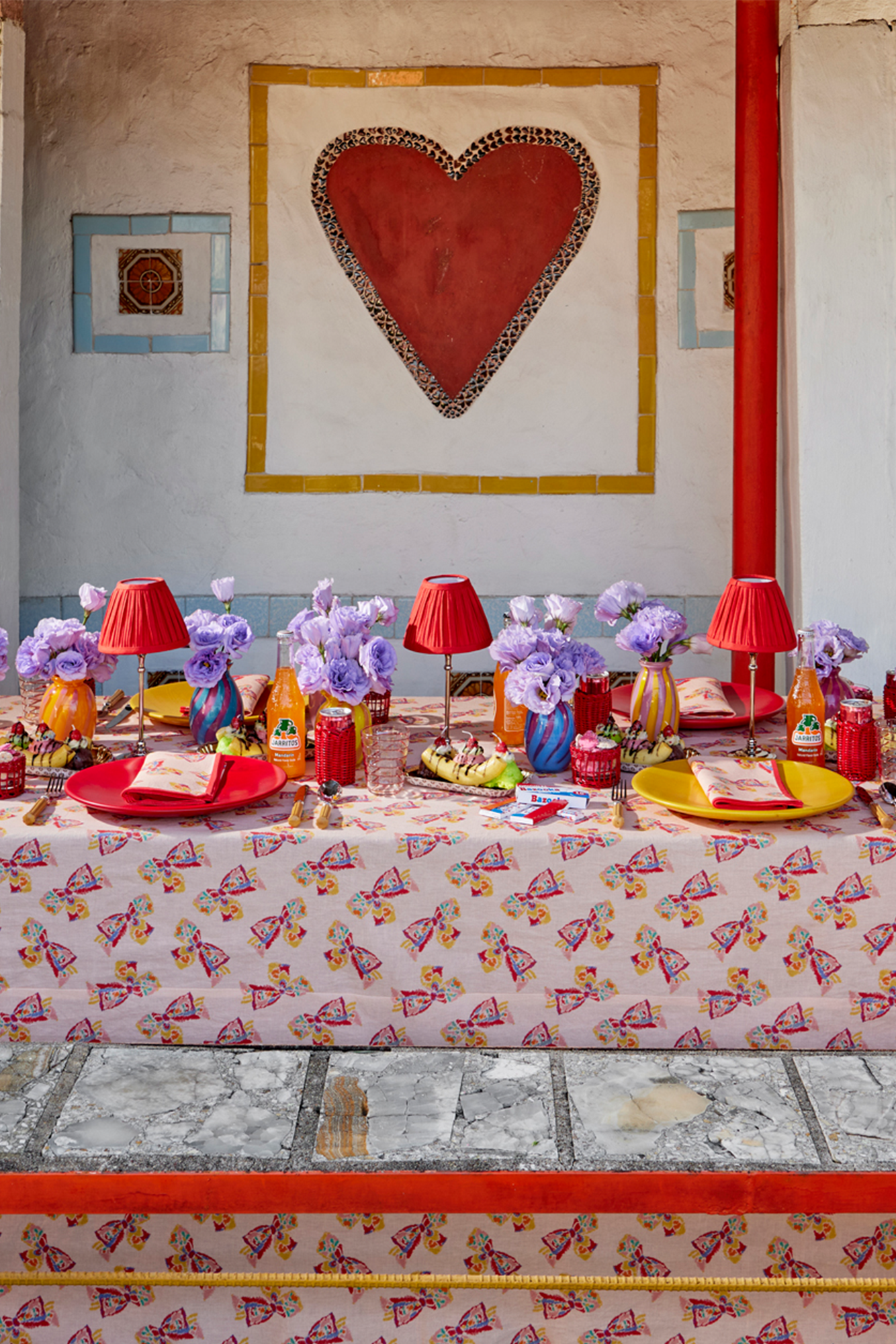 Table setting on the ground featuring a pink tablecloth with a printed pattern of a vintage-inspired rainbow bow. Atop the table are colorful place settings, red shaded table lamps, and fresh purple flowers in blown glass vases.
