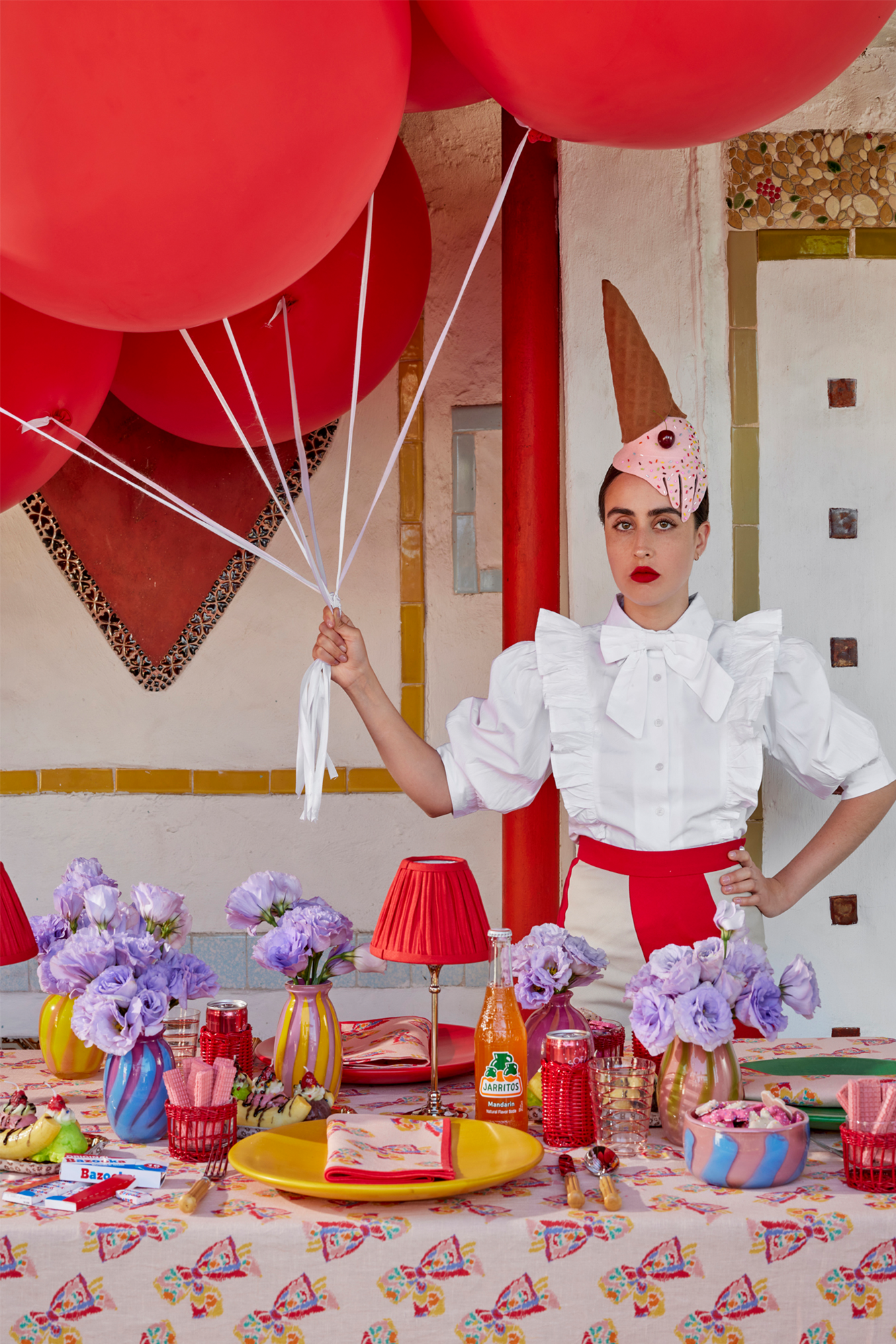 Table setting featuring a pink tablecloth with a printed pattern of a vintage-inspired rainbow bow. Atop the table are colorful place settings, red shaded table lamps, and fresh purple flowers in blown glass vases. Behind the table is a young woman wearing a melting ice cream cone party hat and holding several large red balloons.
