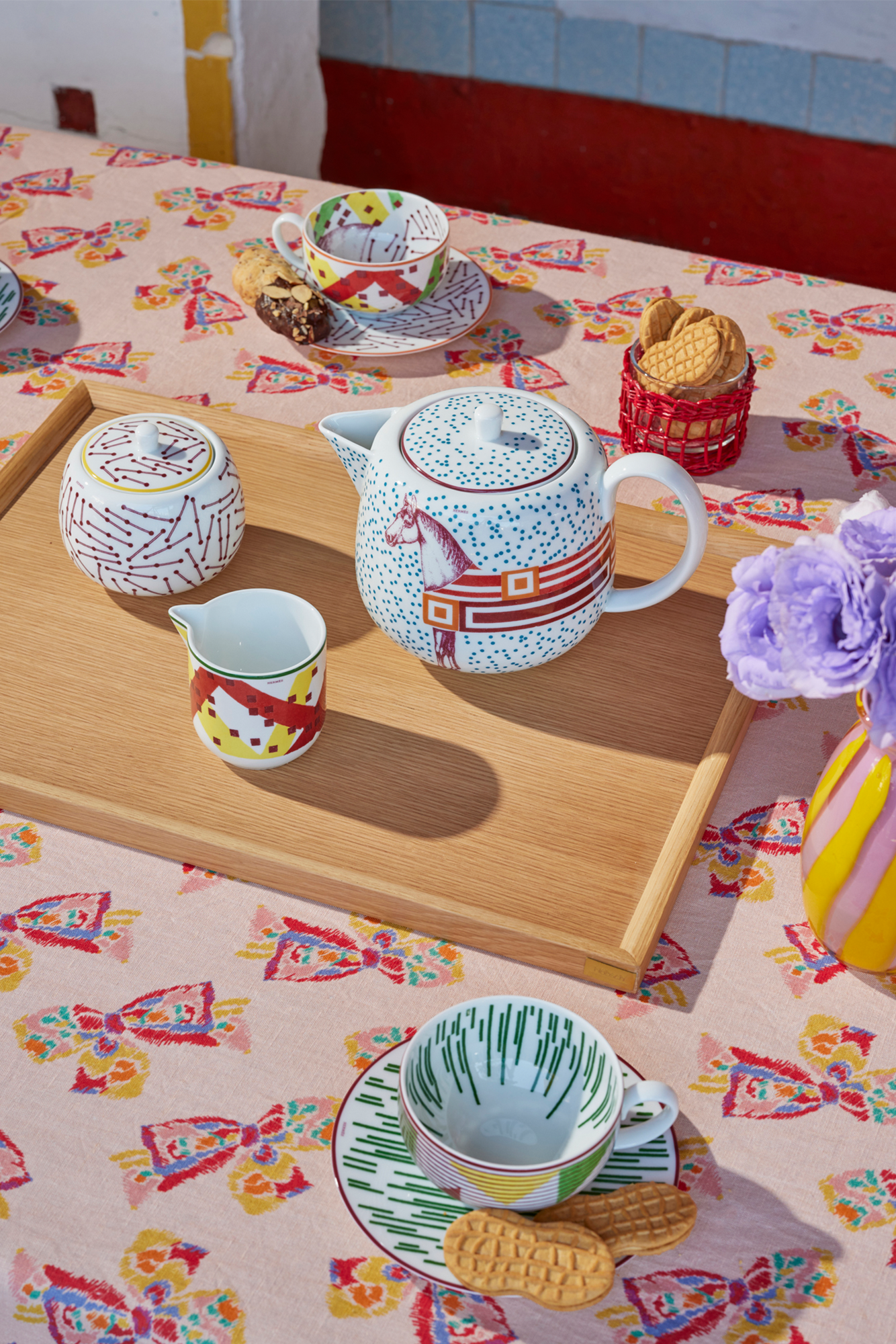 Table setting featuring a pink tablecloth with a printed pattern of a vintage-inspired rainbow bow. Atop the table is an Hermes tea set, resting on a large oak passing tray.