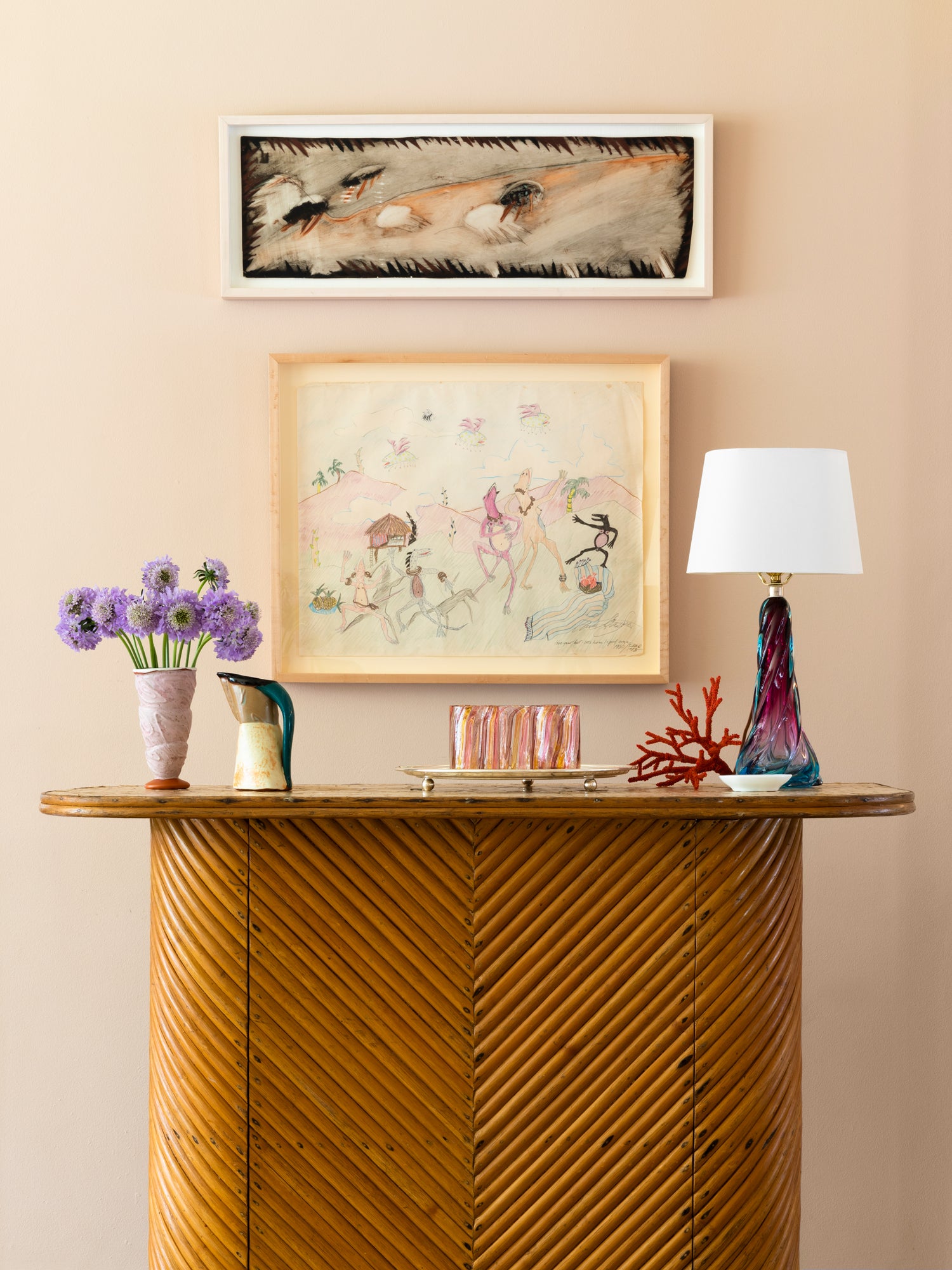 Houses & Parties vignette featuring a striking chevron-patterned wooden console topped with artful curiosities: purple scabiosa blooms, a handcrafted pitcher, a vibrant coral specimen, and a sculptural lamp with purple-blue glass base. Two distinctive framed artworks above—an abstract with textural elements and a whimsical sketch—complete this luxuriously irreverent composition blending natural materials with artistic expression.