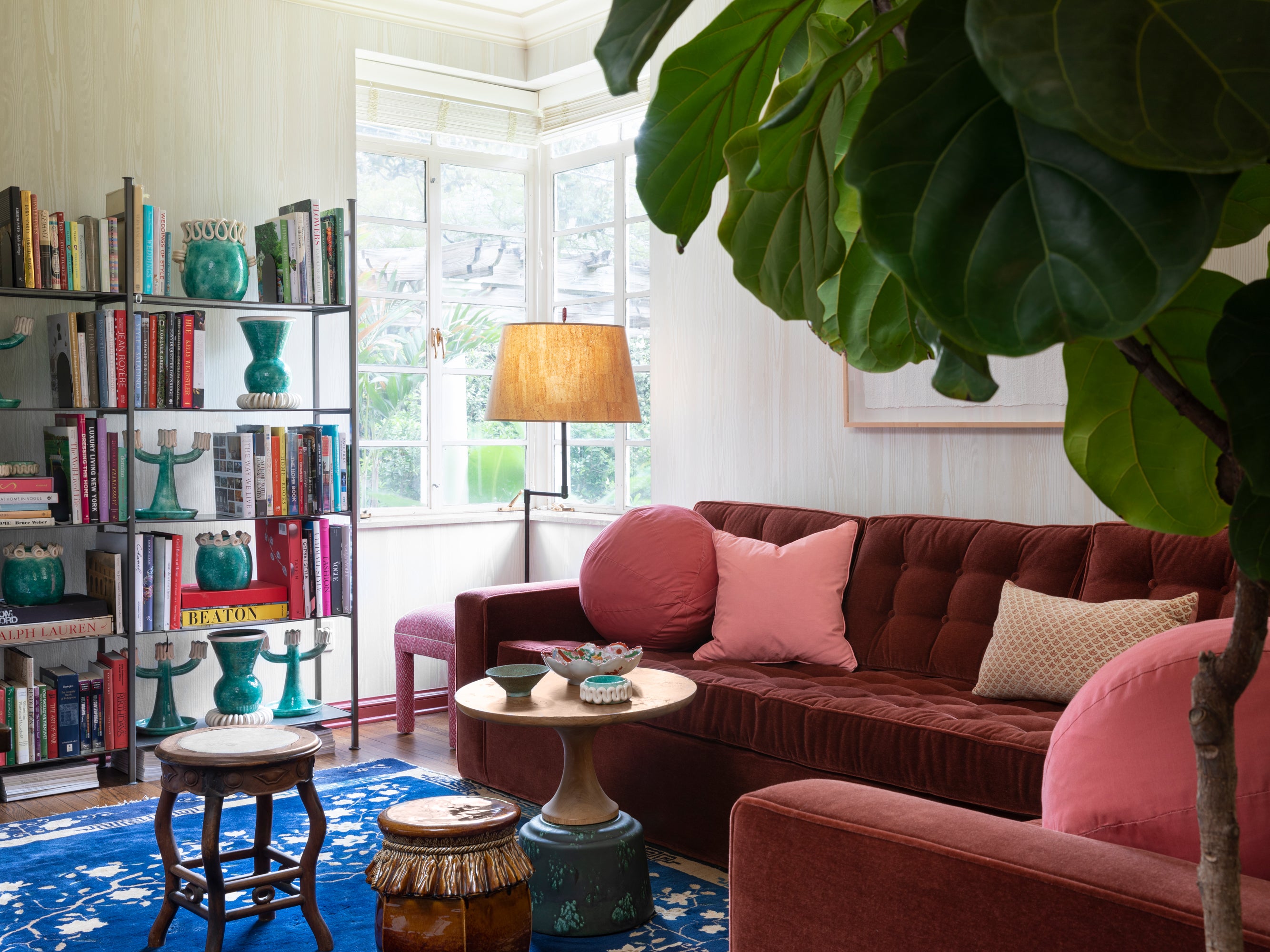 Inviting library nook featuring a rich chocolate velvet tufted sofa with coral accent pillows, surrounded by colorful ceramic vessels, open bookshelves, and a statement fiddle leaf fig plant – demonstrating Houses & Parties' skill at creating livable, collected spaces that balance masculine and feminine elements through color and texture.