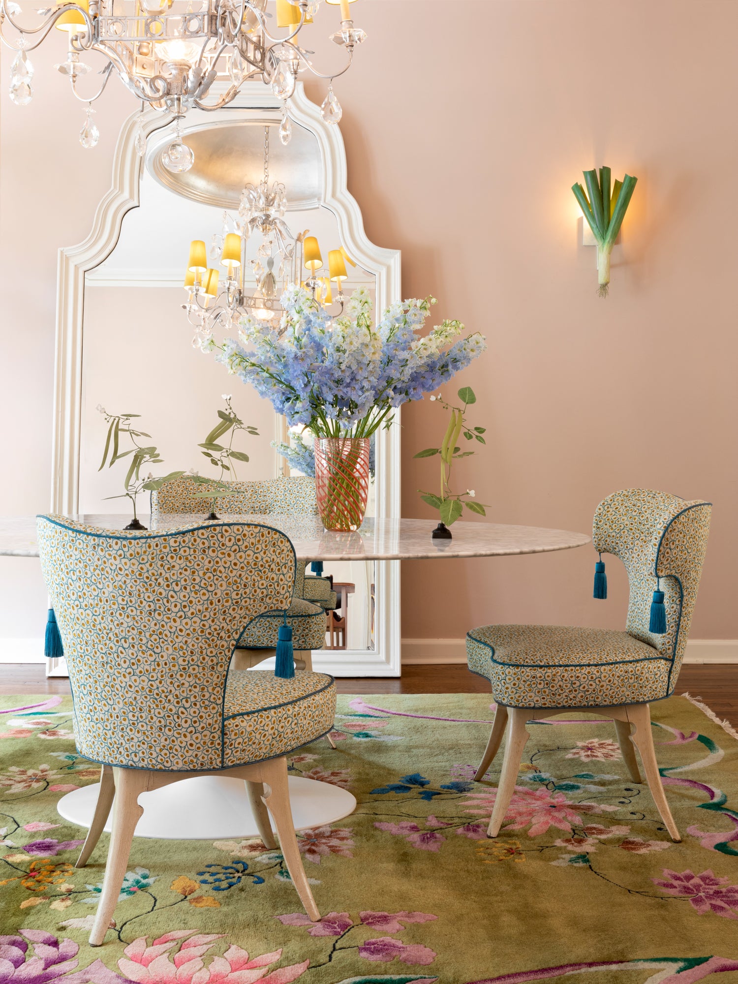 Houses & Parties dining space featuring custom chairs upholstered in blue-and-gold leopard print fabric with playful tassels. A round marble table showcases vibrant blue delphinium blooms in a glass vase. An ornate white mirror frames the scene beneath a crystal chandelier with yellow shades, while a whimsical leek-shaped sconce provides unexpected humor. The floral carpet in soft greens, pinks, and blues completes this luxuriously irreverent setting that perfectly balances sophistication with playfulness.