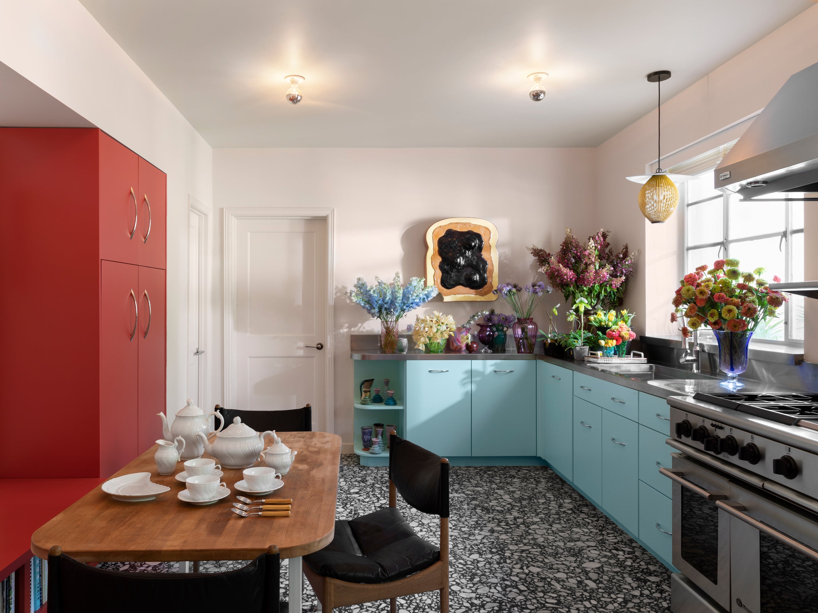 Houses & Parties kitchen combining playful color with practical luxury—featuring robin's egg blue cabinetry alongside coral storage units. The countertop overflows with vibrant fresh flowers in colorful vessels, while a wooden table set with white porcelain tea service creates an inviting dining spot.