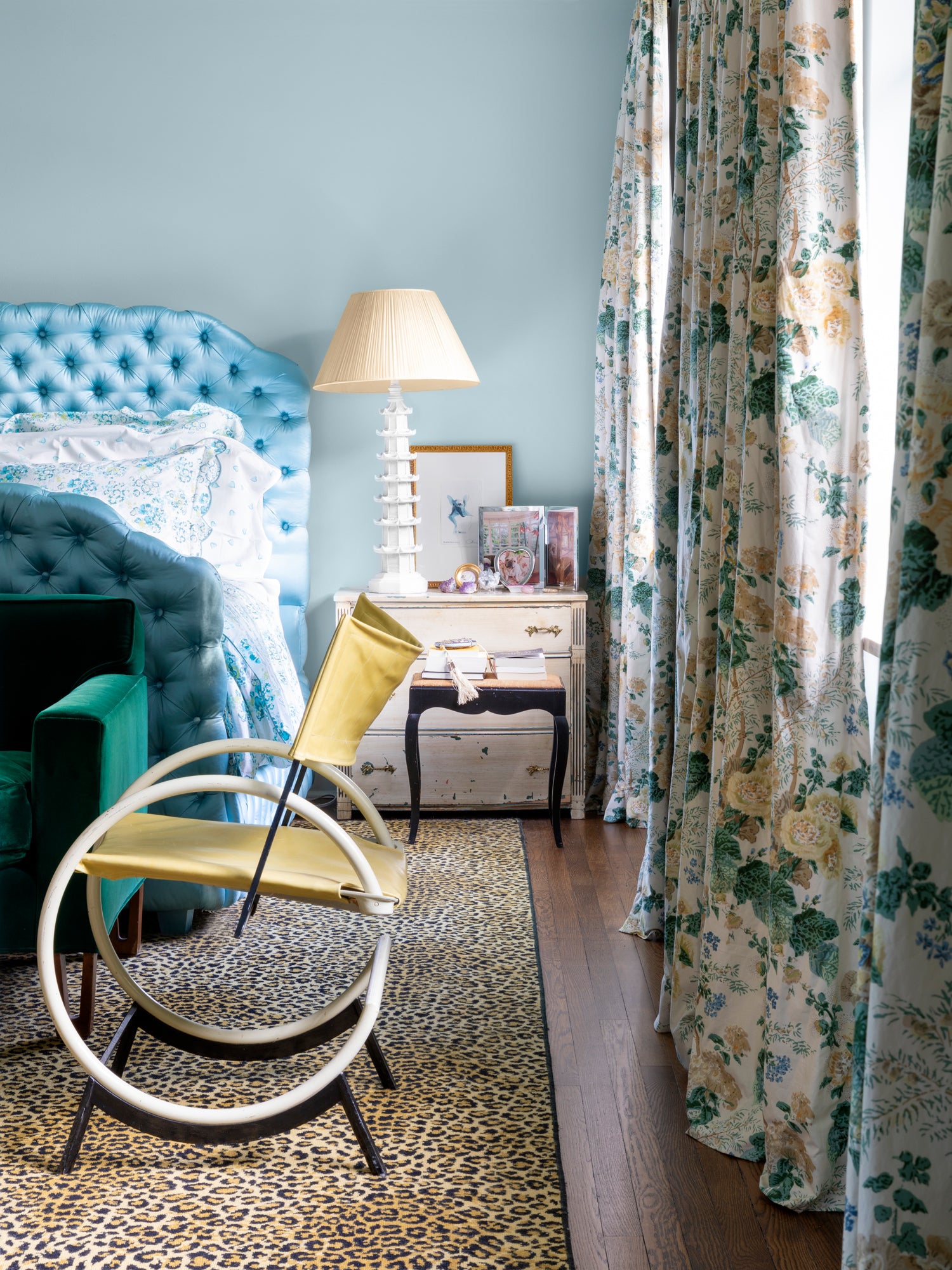 Eclectic bedroom featuring a sky-blue tufted headboard, leopard print area rug, modernist yellow slingback chair, and floral draperies – exemplifying Houses & Parties' talent for unexpected color combinations and confident pattern mixing that creates spaces both sophisticated and full of personality.