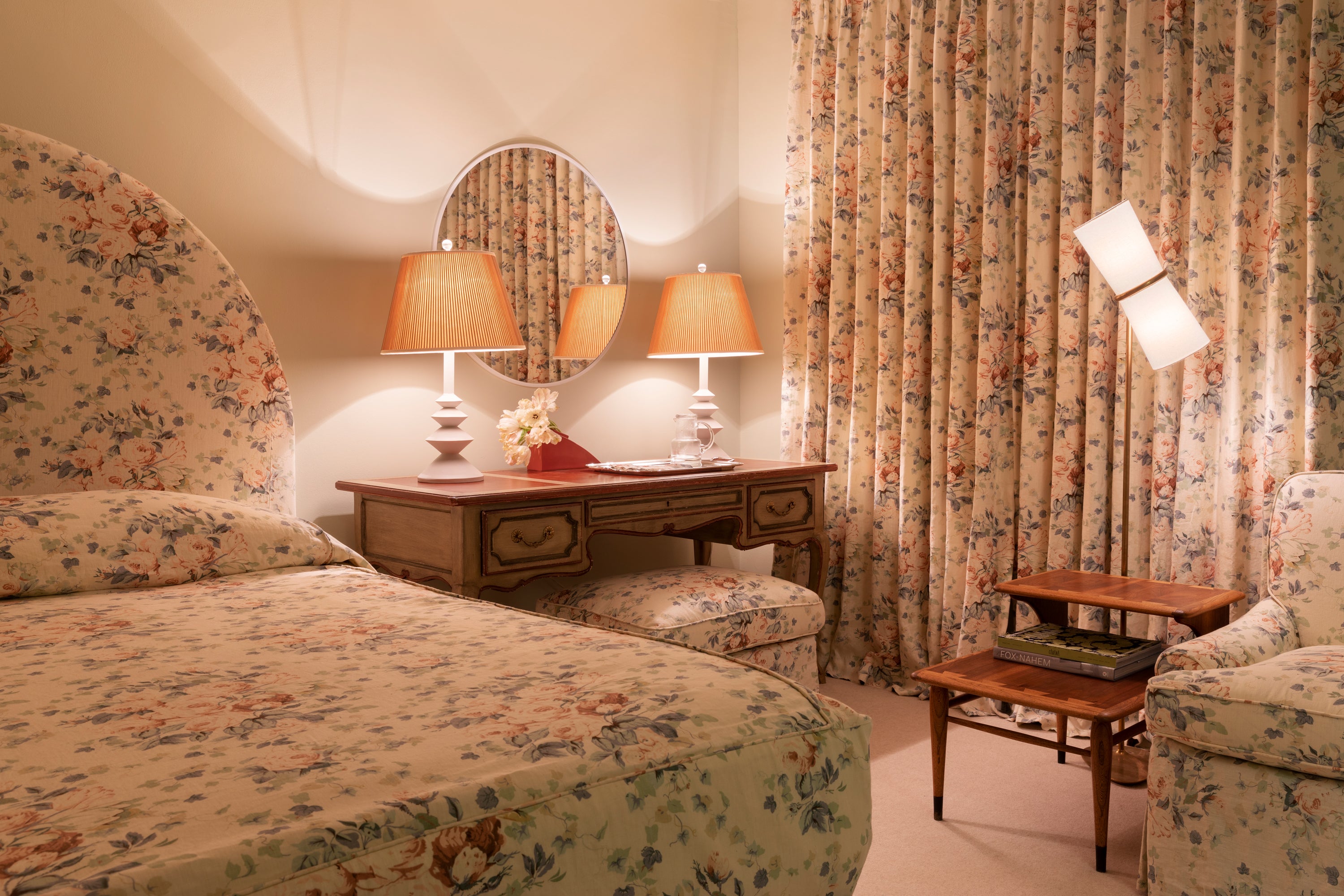 Houses & Parties bedroom featuring coordinated floral patterns on bedding, curtains, and upholstery in soft peach and blue tones. A traditional wooden desk with stacked ceramic lamps, oval mirror, and red accent vase creates a focal point between bed and window. A modern wall sconce adds unexpected contrast to this luxurious space that balances vintage romance with contemporary touches.