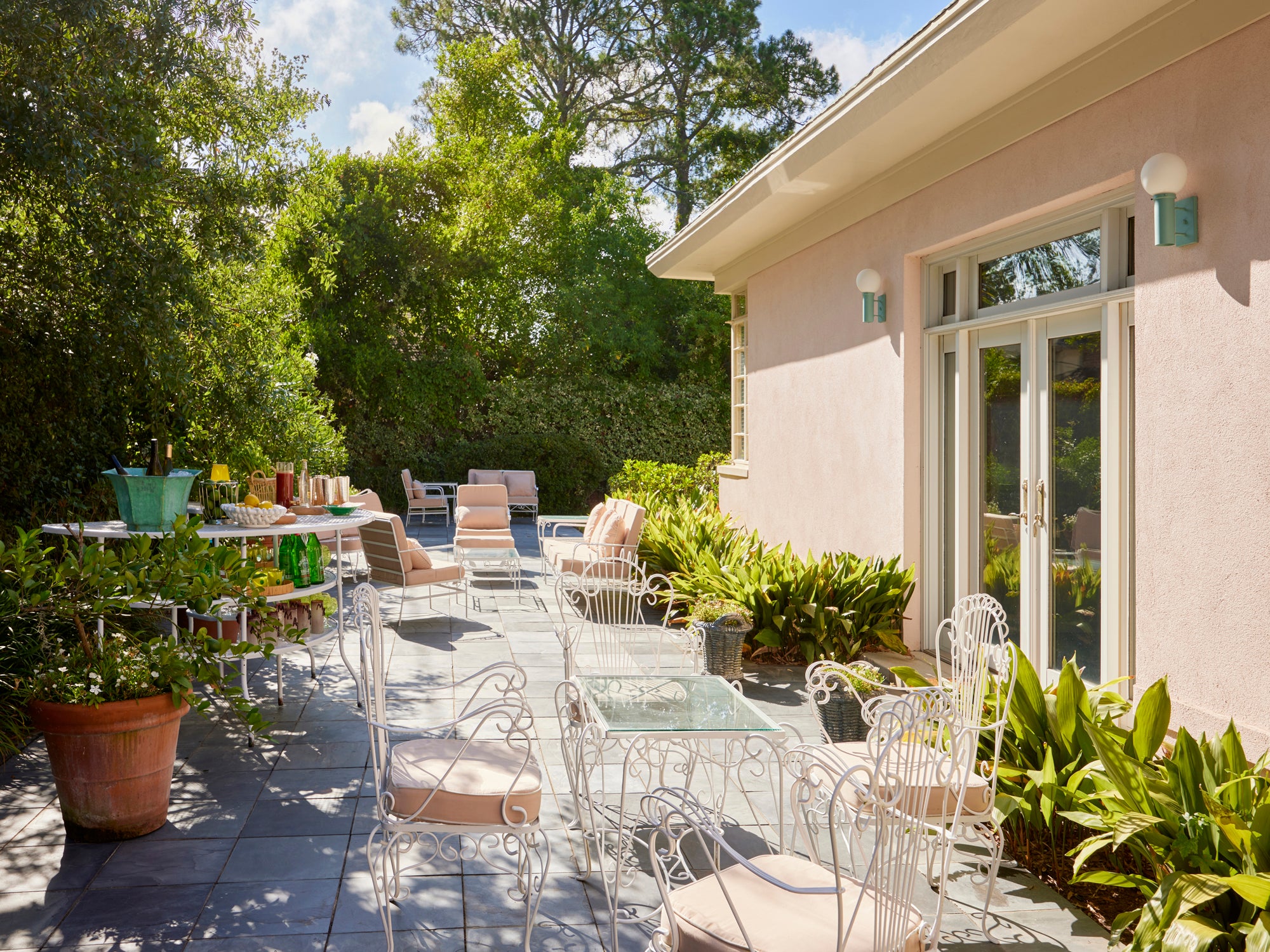 Houses & Parties garden terrace featuring vintage white wrought-iron furniture with blush cushions arranged for elegant outdoor entertaining. A bar cart displays green glassware and cocktail accessories while potted plants provide lush greenery. Mint-colored sconces accent the pale stucco exterior with French doors opening to the patio. Surrounded by mature trees creating natural privacy, this sophisticated outdoor space balances classic garden elements with contemporary comforts.
