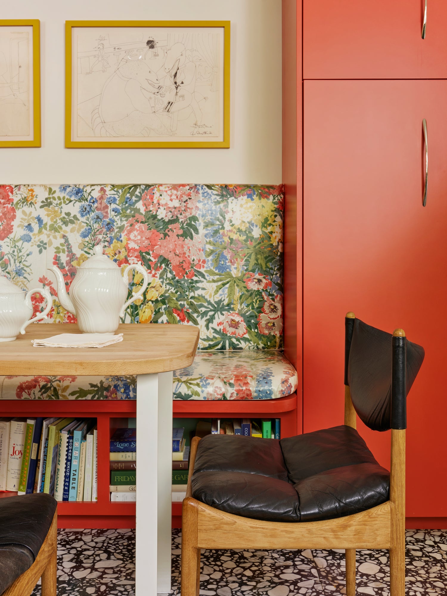 Houses & Parties breakfast nook featuring a vibrant coral-painted built-in banquette upholstered in lush floral fabric with bookshelf below. A light wood table paired with minimalist white ceramics creates contrast against the colorful backdrop. A mid-century Danish chair with black leather cushions adds architectural interest, while yellow-framed sketches hang above. The terrazzo flooring ties together this irreverent color story—blending vintage elements with bold modern choices.