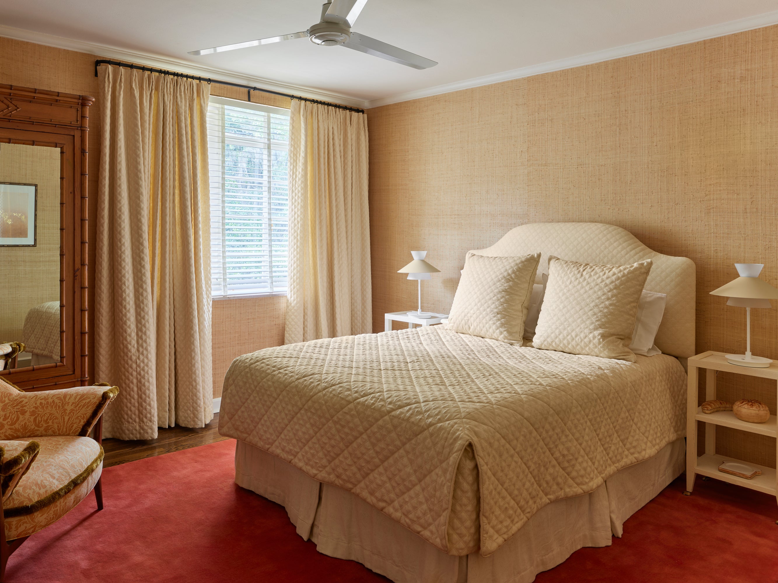Monochromatic bedroom featuring subtle textural interplay in warm ivory and cream tones against grasscloth wallpaper. A quilted bedspread and matching pillows complement an upholstered headboard, while cream curtains filter light by the window. Modern white table lamps flank the bed, with a vintage faux-bamboo mirror bringing architectural interest. The bold coral-red carpet provides unexpected contrast, demonstrating the brand's signature ability to balance tranquil elegance with daring color accents.
