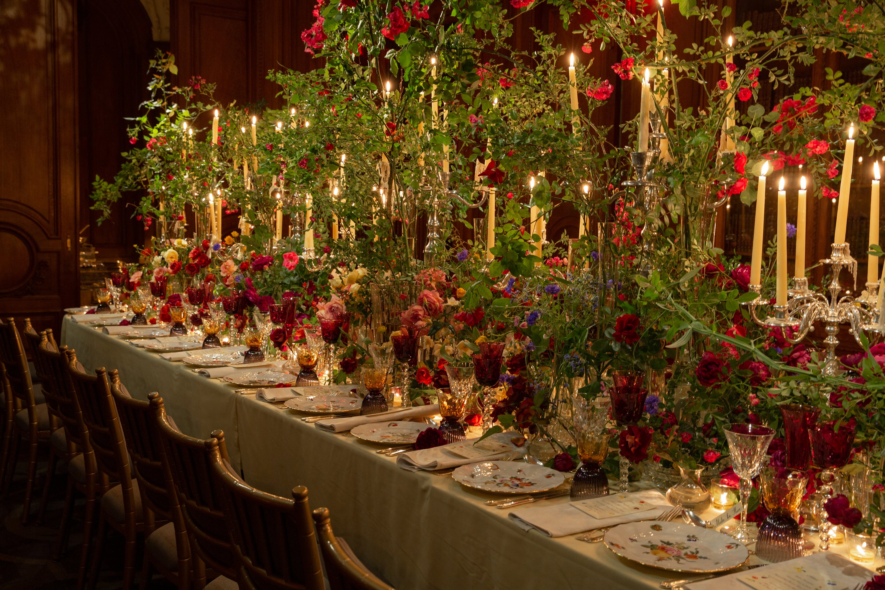 Breathtaking formal dining table adorned with an enchanted garden-inspired tablescape featuring cascading red flowering branches, crystal candelabras with tall tapers, and jewel-toned glassware – all bathed in warm candlelight for an opulent, romantic celebration in a wood-paneled estate setting.