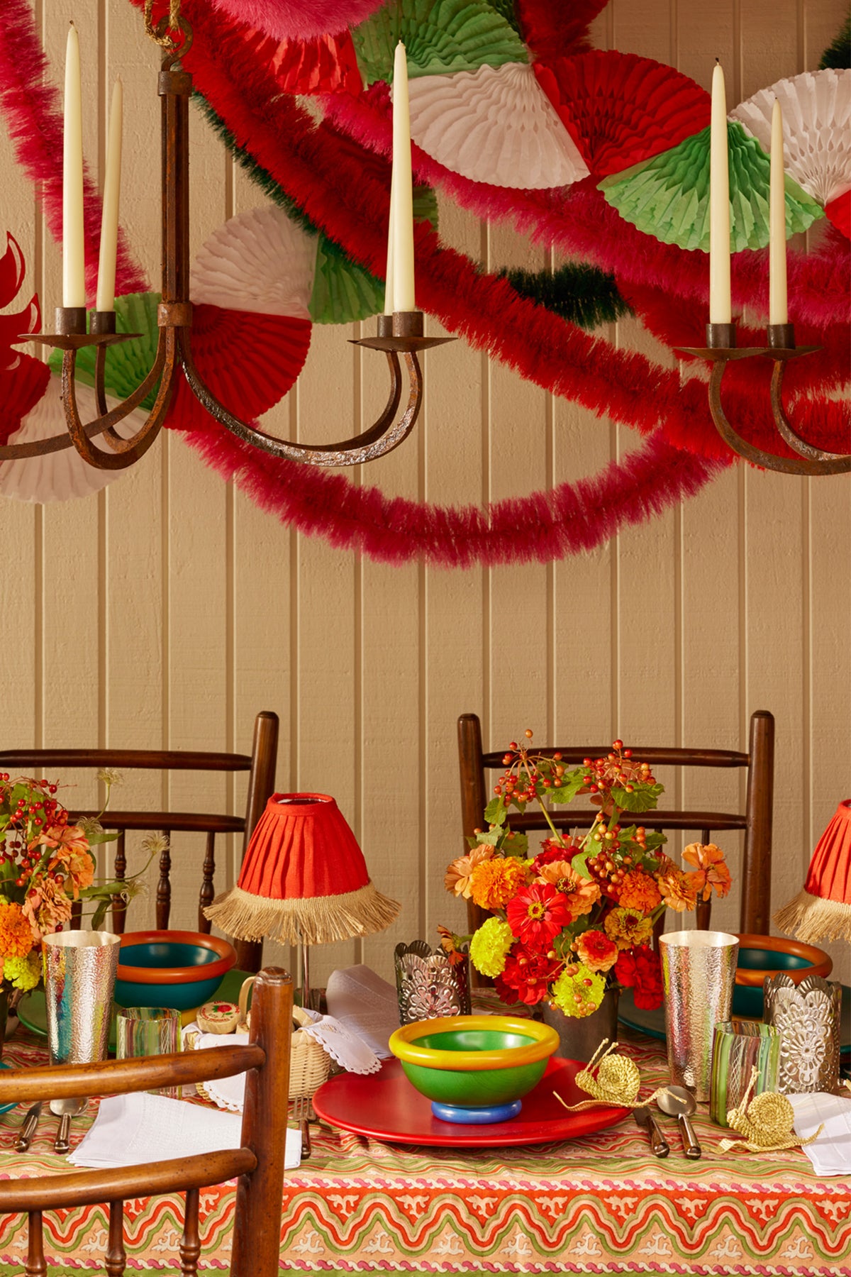 Table setting underneath iron chandeliers draped in red, white, and green paper garland. Table covered in a patterned tablecloth and set with red plates, multicolor bowls, silver tumblers, table lamp with red lampshade with tan fringe, fresh flowers, and corn husk snails trailing across the tablescape.