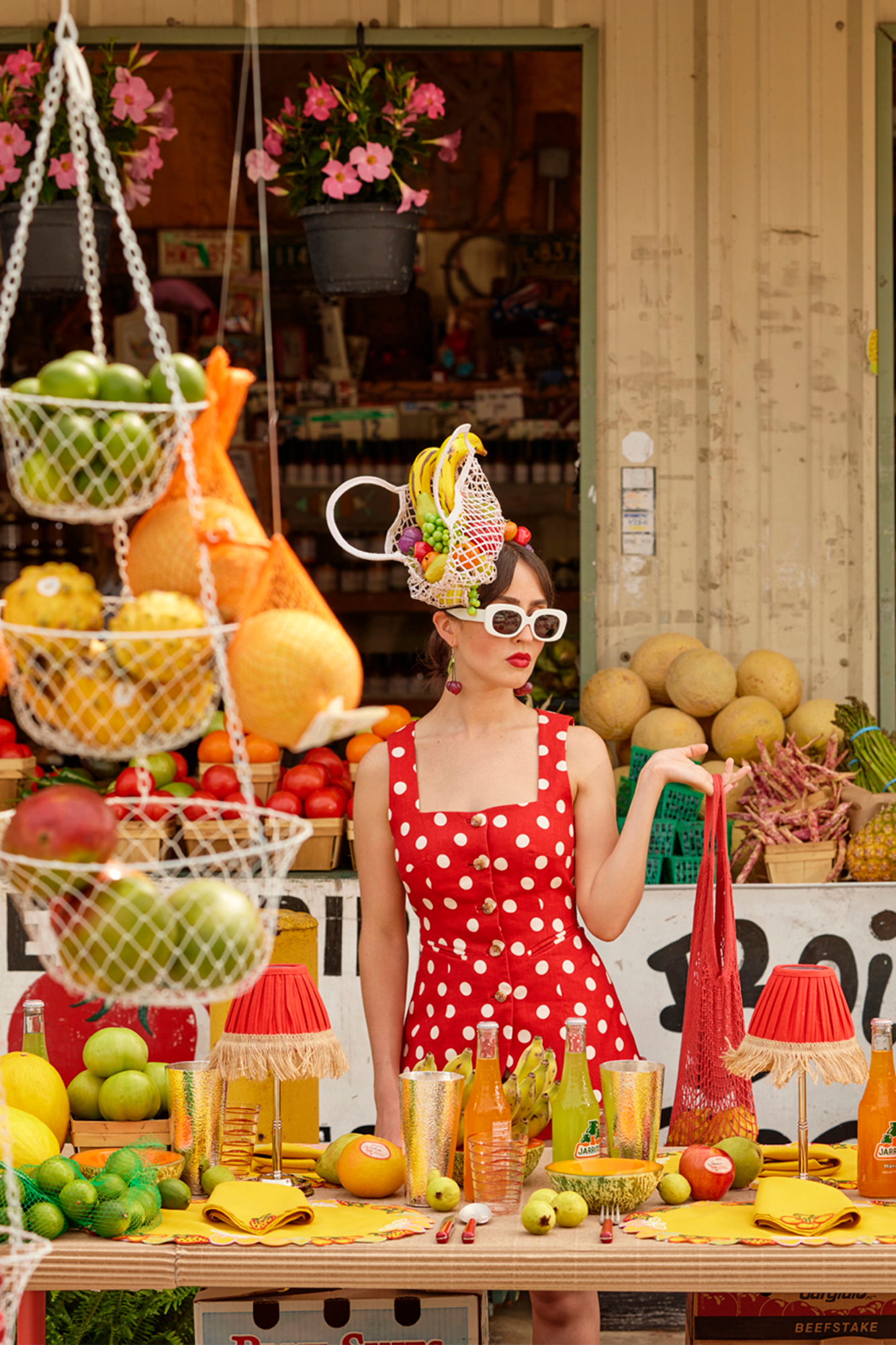 Woman in front of tabletop at a fruit market with a fruit bag hat, picante fringe lamps, hammered silver tumblers, colorful fruit placemats and napkins, and a Beechwood serving bowl on a coordinated tripod.